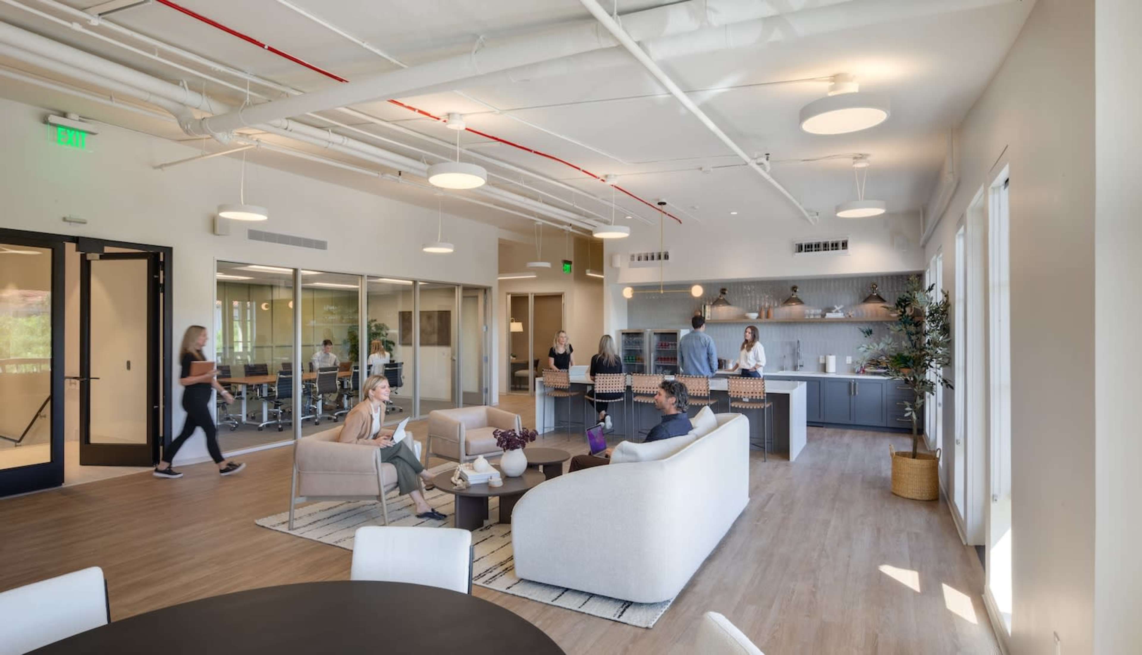 A modern office lounge area with people engaging in conversations, a kitchenette in the background, and large windows allowing natural light.