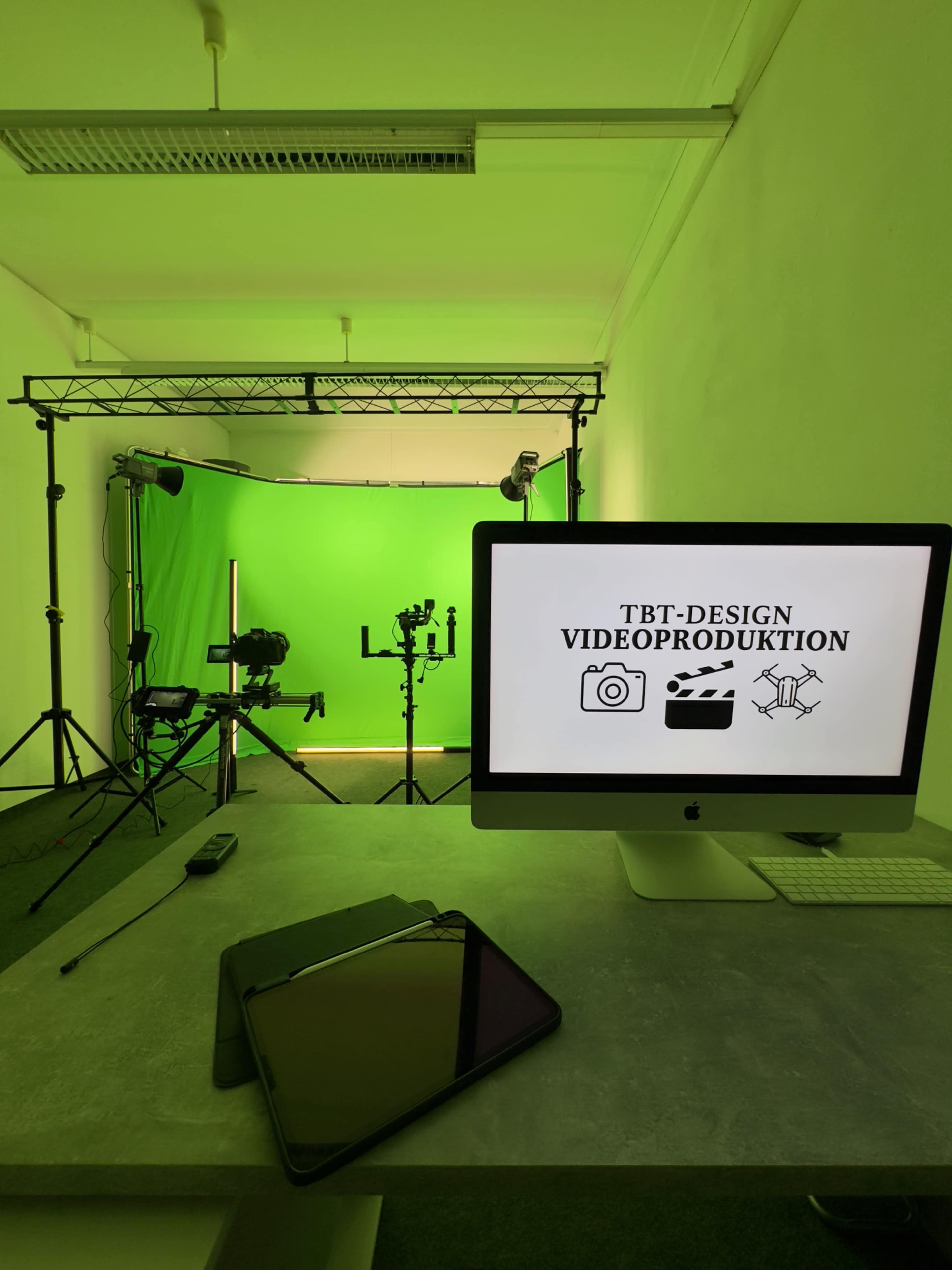 The image shows a video production studio with a green backdrop, lighting equipment, and a computer displaying a logo on a desk.