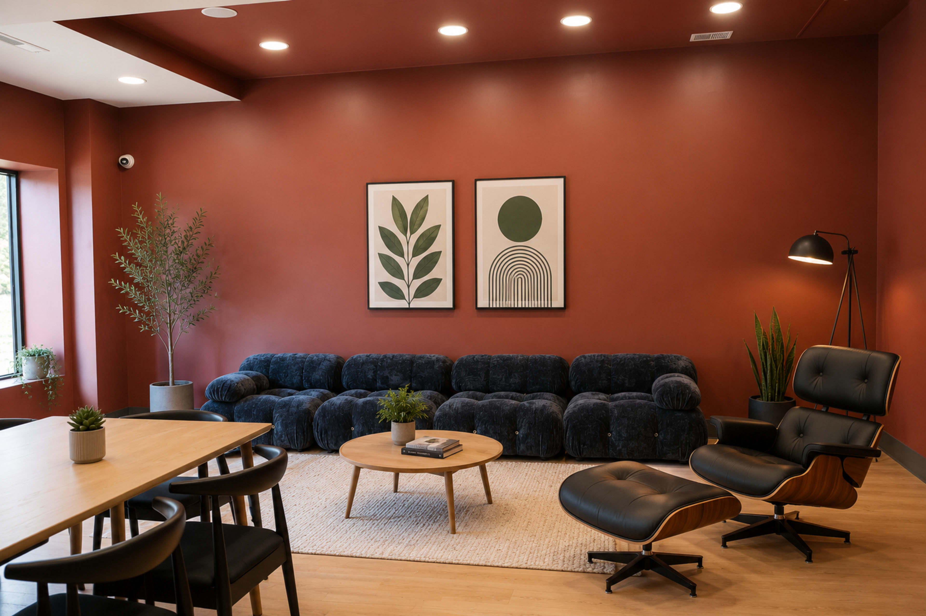 The image shows a modern lounge area with a dark blue sectional sofa, a round wooden coffee table, and a dining table, all against a red wall featuring two framed art pieces.