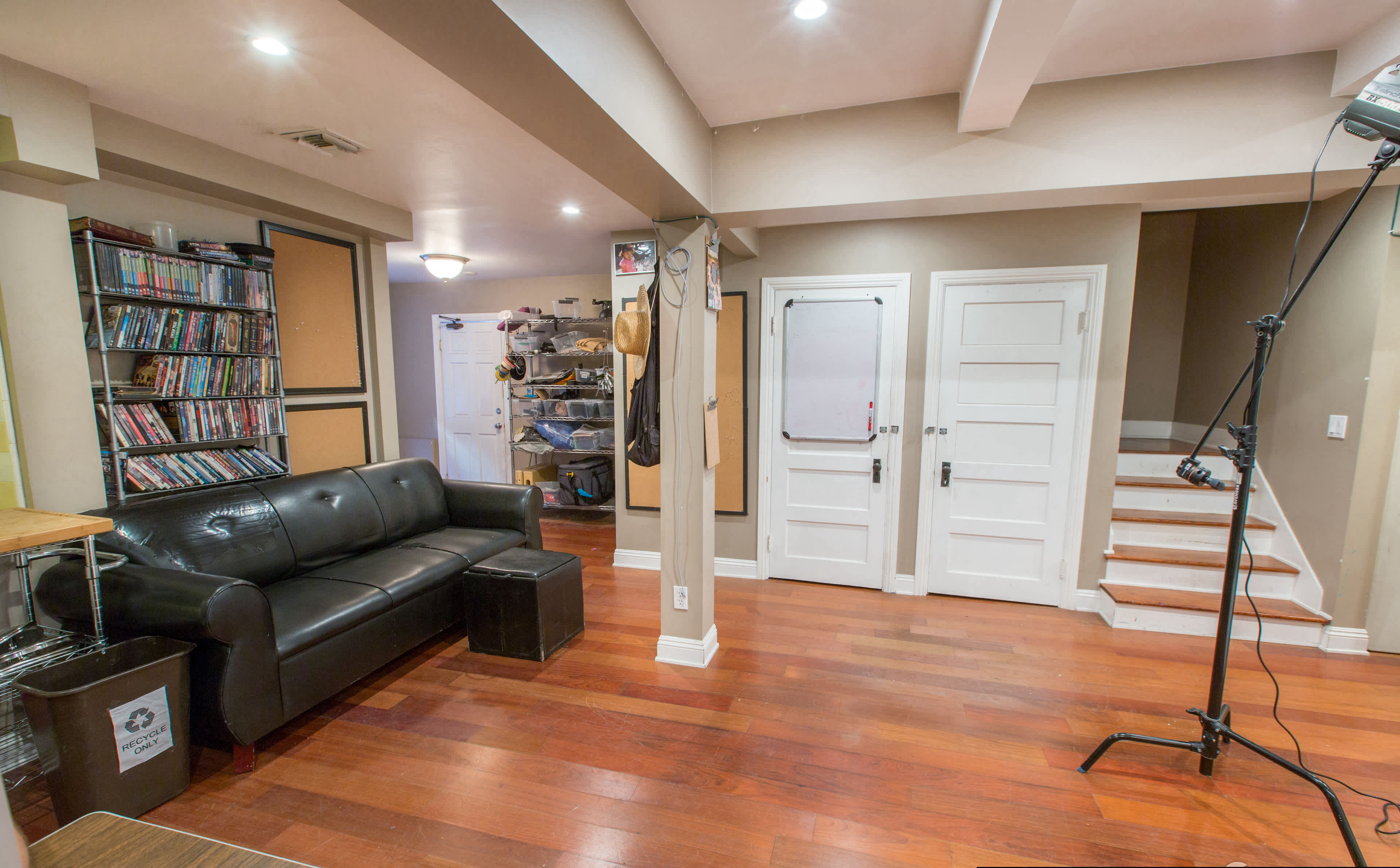 A living space with a black couch, a wooden table, a bookshelf filled with DVDs, and stairs leading to an upper level.