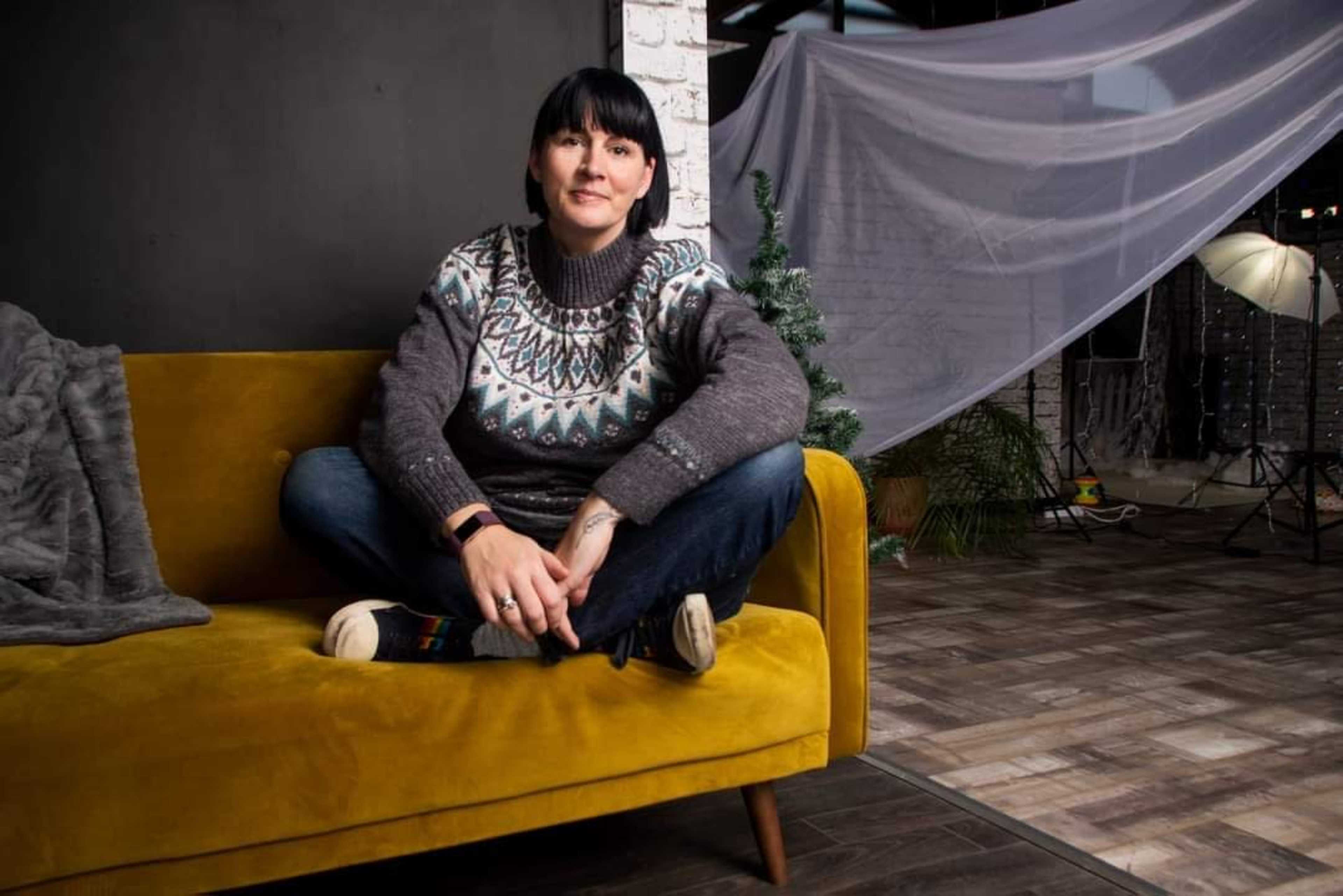 A person sits cross-legged on a mustard-yellow couch in a studio with a backdrop and lighting equipment.