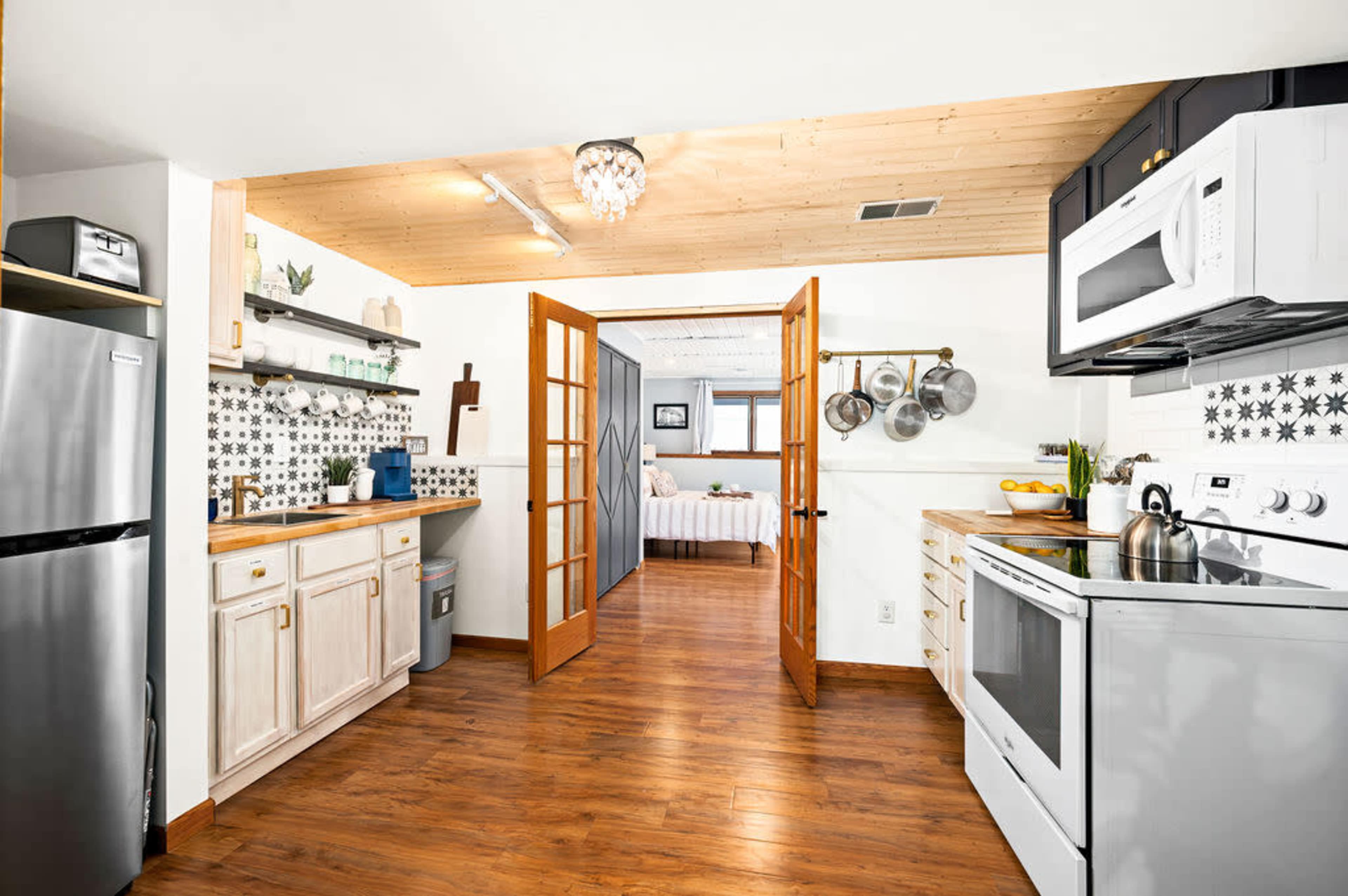 The image shows a modern kitchen with wooden cabinetry, stainless steel appliances, and a doorway leading to a bright, open living space with a bed in the background.
