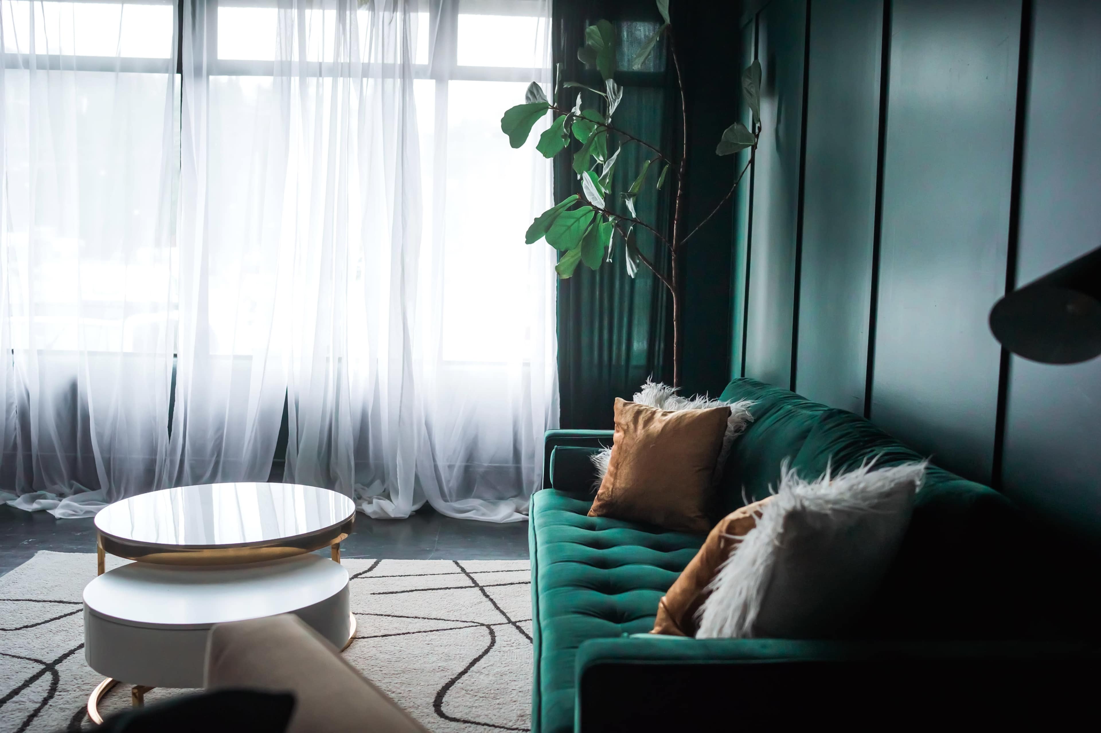 A green velvet sofa with decorative pillows sits opposite a round coffee table in a well-lit living room with sheer curtains.
