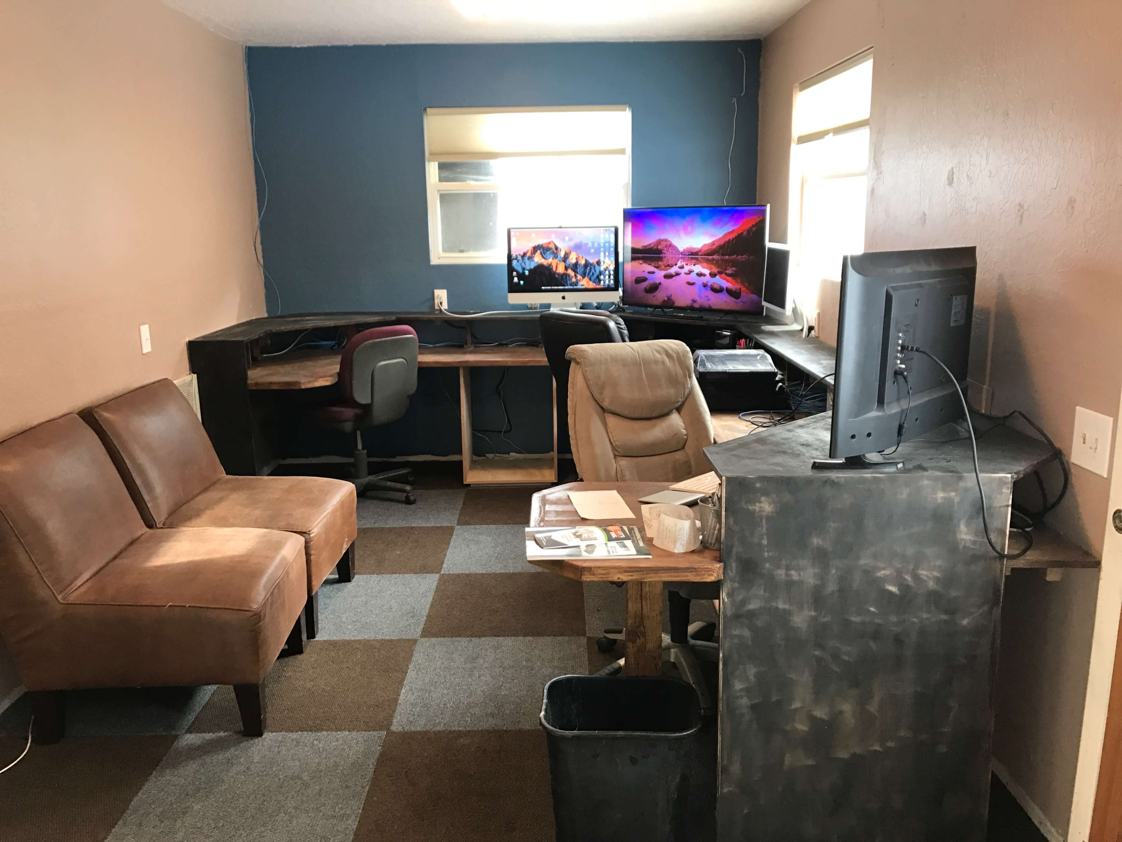 The image shows a small office space with two computer desks, a brown couch, and two monitors mounted on the wall.