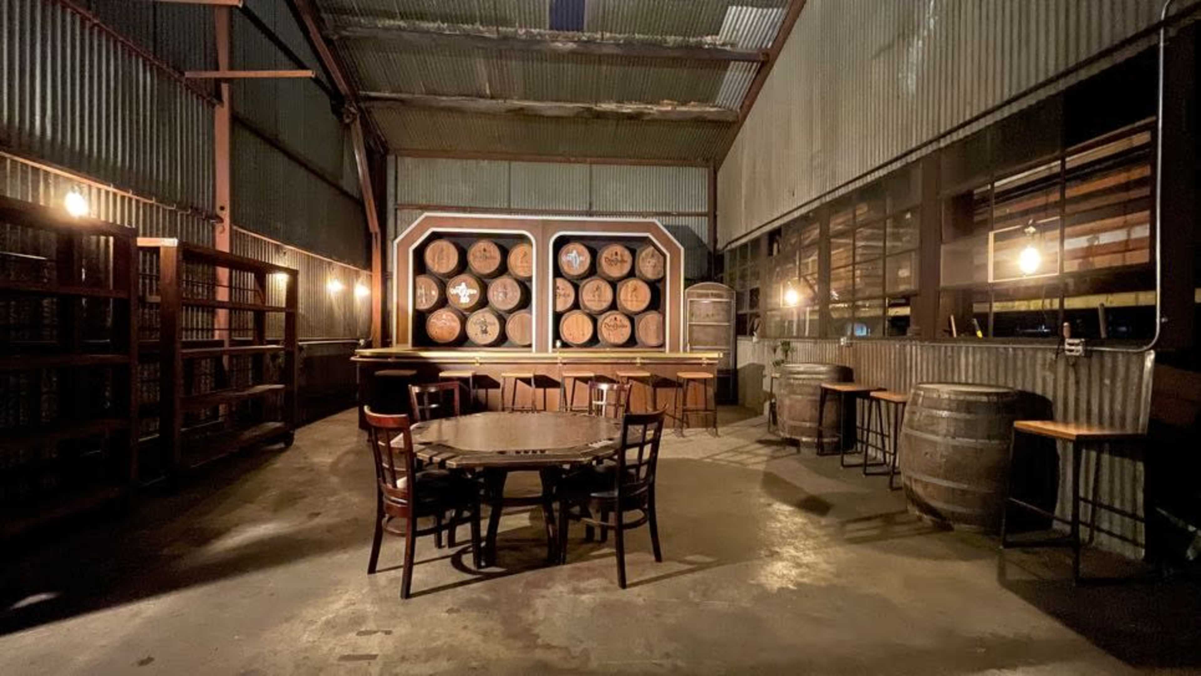 The image shows a rustic interior space with wooden barrels, a large display of barrels on the wall, and several tables and chairs arranged in a dimly lit setting.