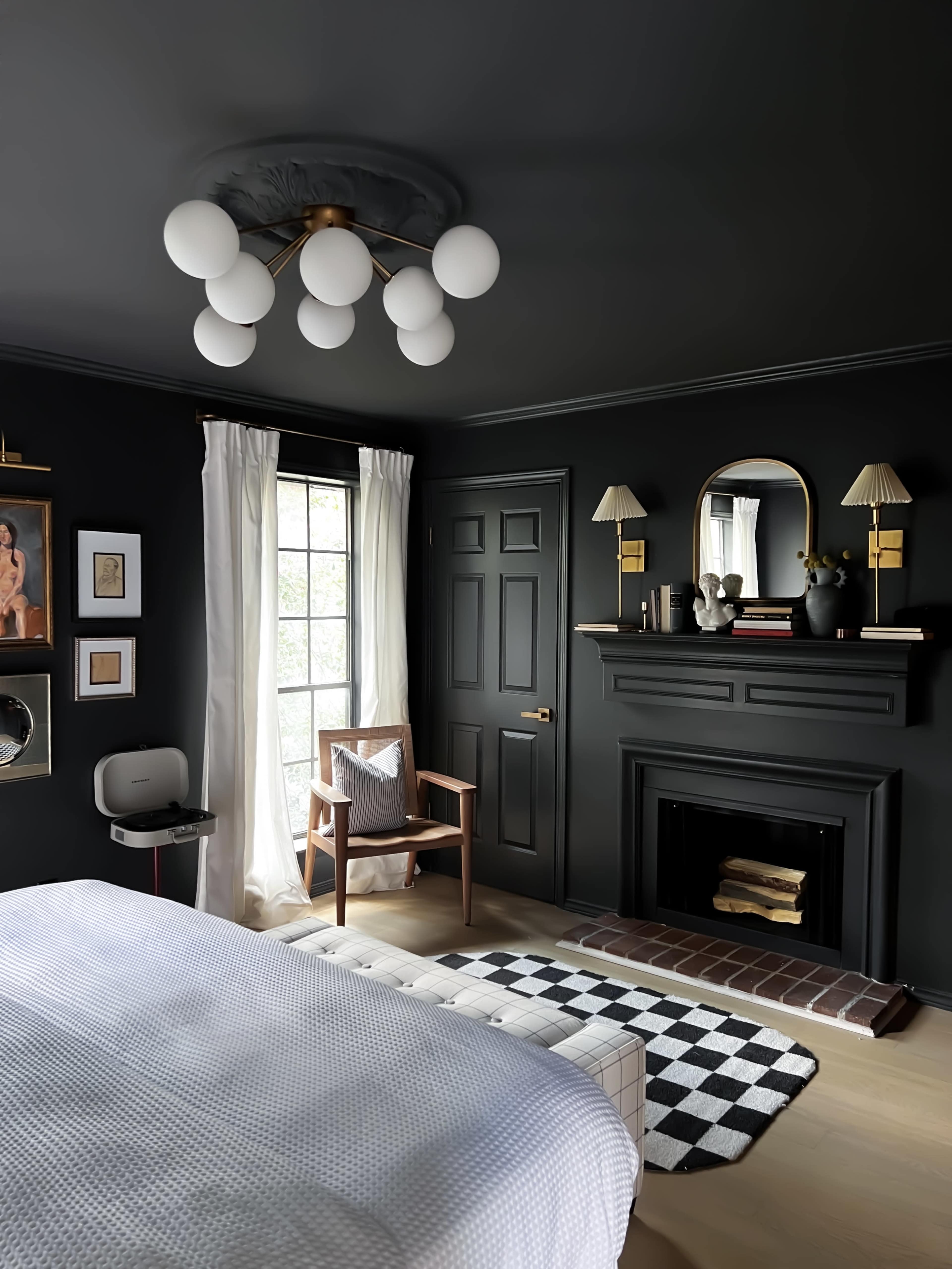 The image shows a modern bedroom with dark walls, a fireplace, large round light fixtures, and a combination of decorative elements including framed artwork and a checkered rug.