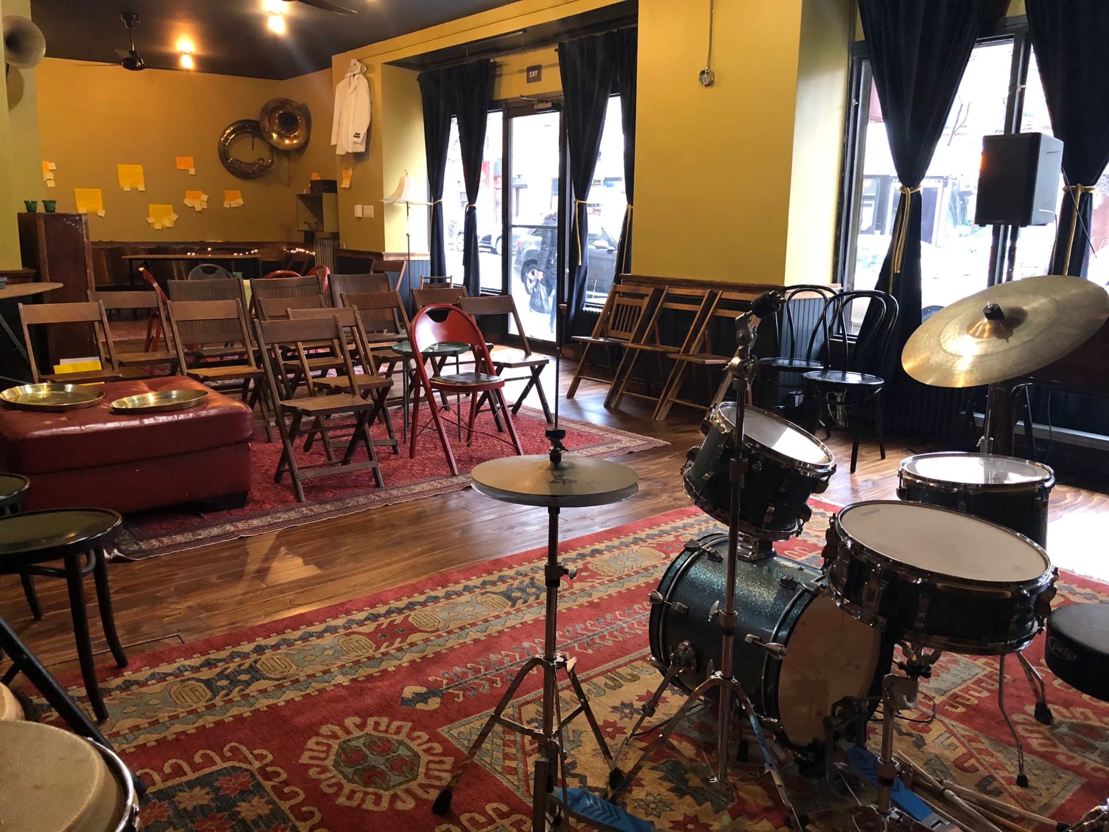 A small performance space features rows of chairs, a drum set, and decorative rugs on a wooden floor, with large windows allowing natural light to enter.