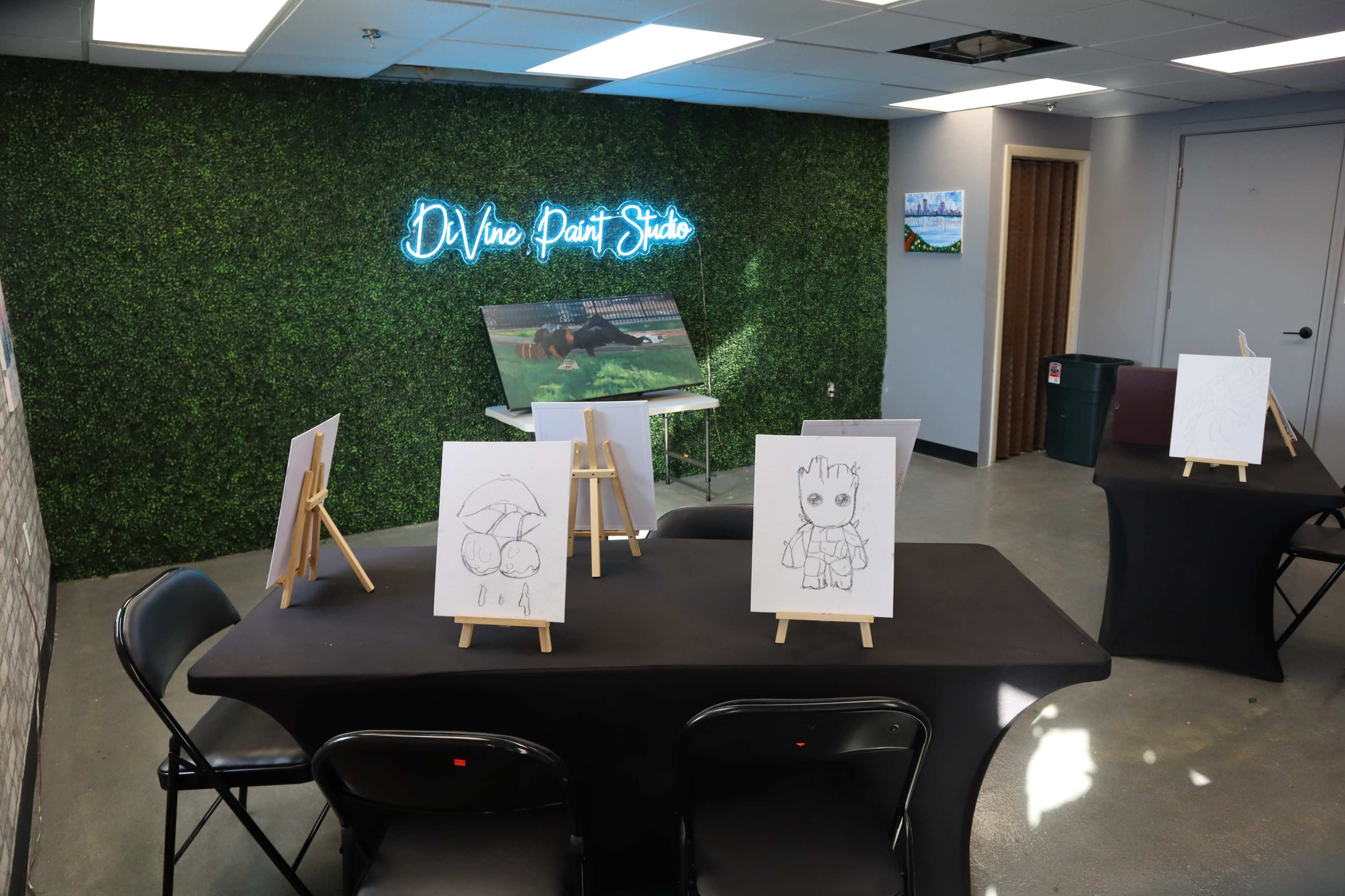 A room labeled "Divine Paint Studio," featuring a green wall with neon signage, a television, and several easels with drawings on display arranged around black tables.
