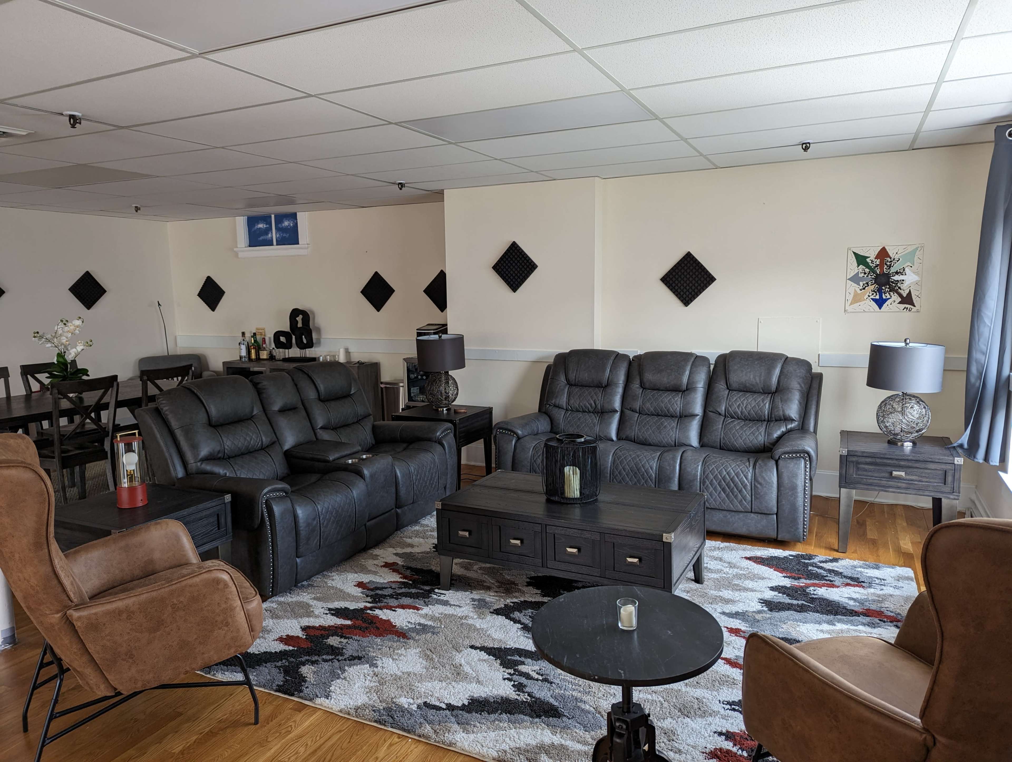 A spacious living room with black leather seating, a coffee table, and a patterned rug, illuminated by natural light through the windows.
