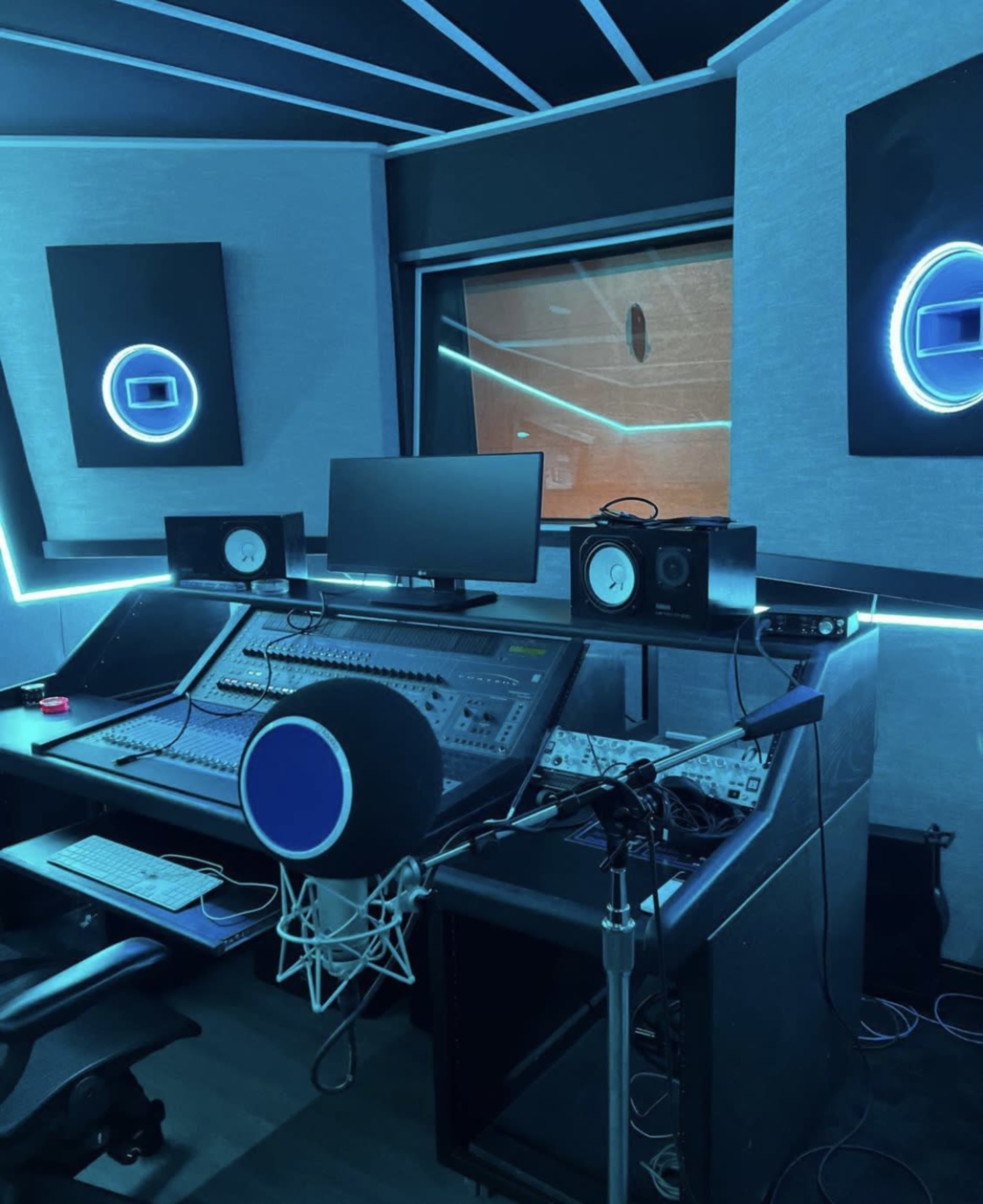 The image shows a modern recording studio with a mixing console, monitor screens, and a microphone setup, illuminated by blue LED lighting.