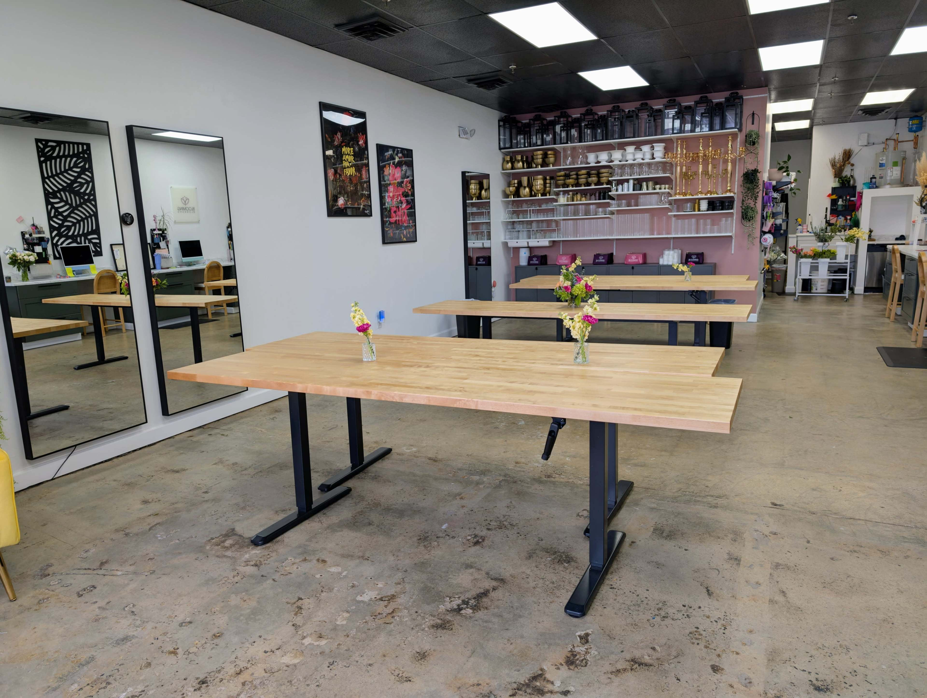 The image shows a bright, modern workspace with multiple wooden tables set up and small flower vases on each table, surrounded by mirrors and a shelf filled with various items.