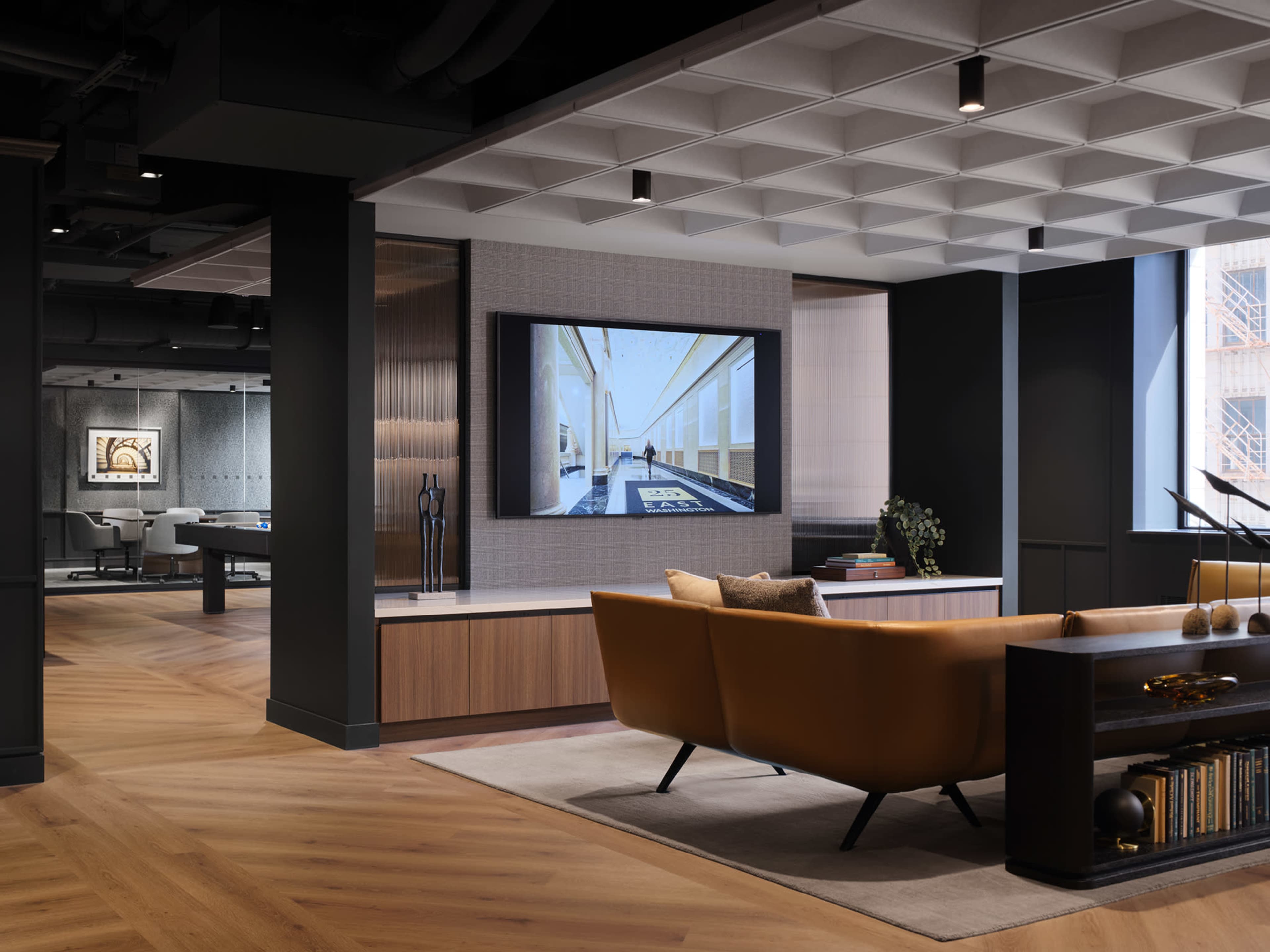 The image shows a modern office lounge area featuring a large television mounted on the wall, a stylish sofa, and wooden flooring with a geometric ceiling design.