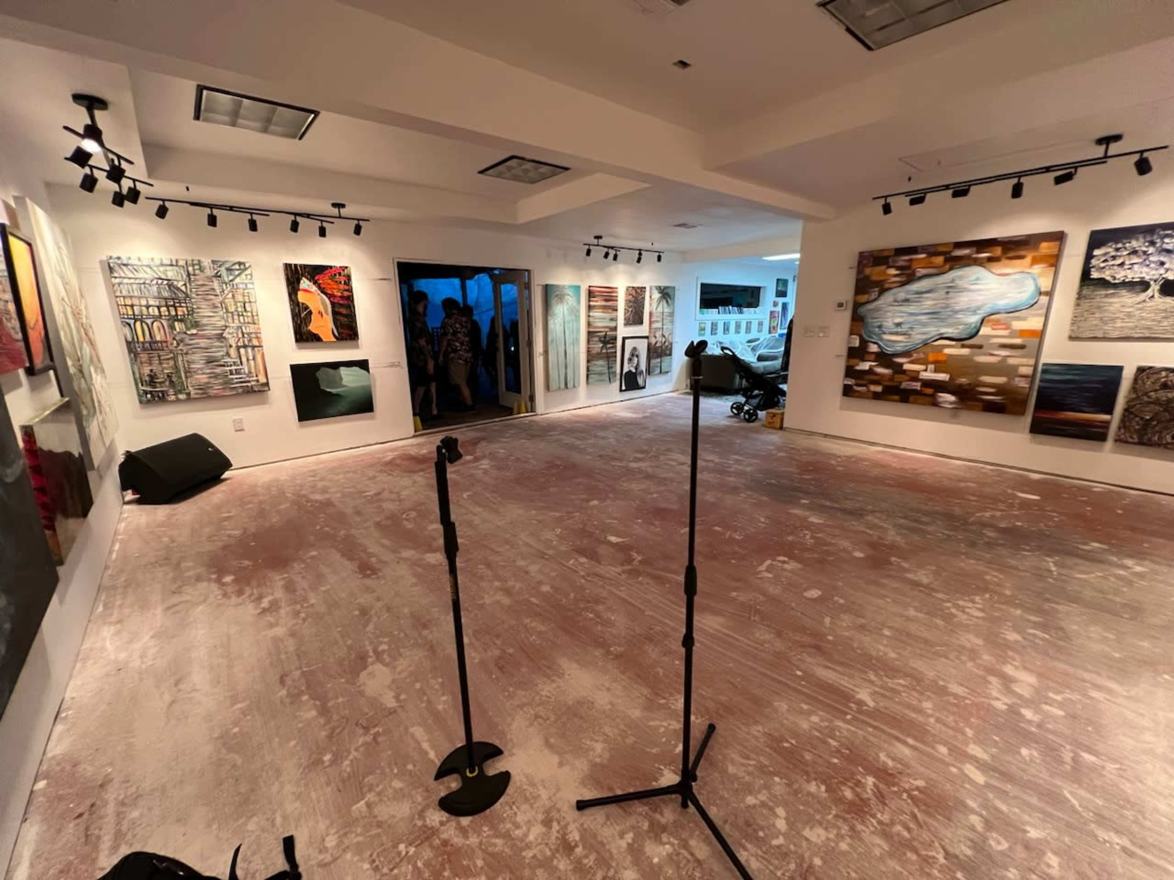 An art gallery with various paintings displayed on the walls and two microphone stands positioned in the center of a vacant, unfinished floor.