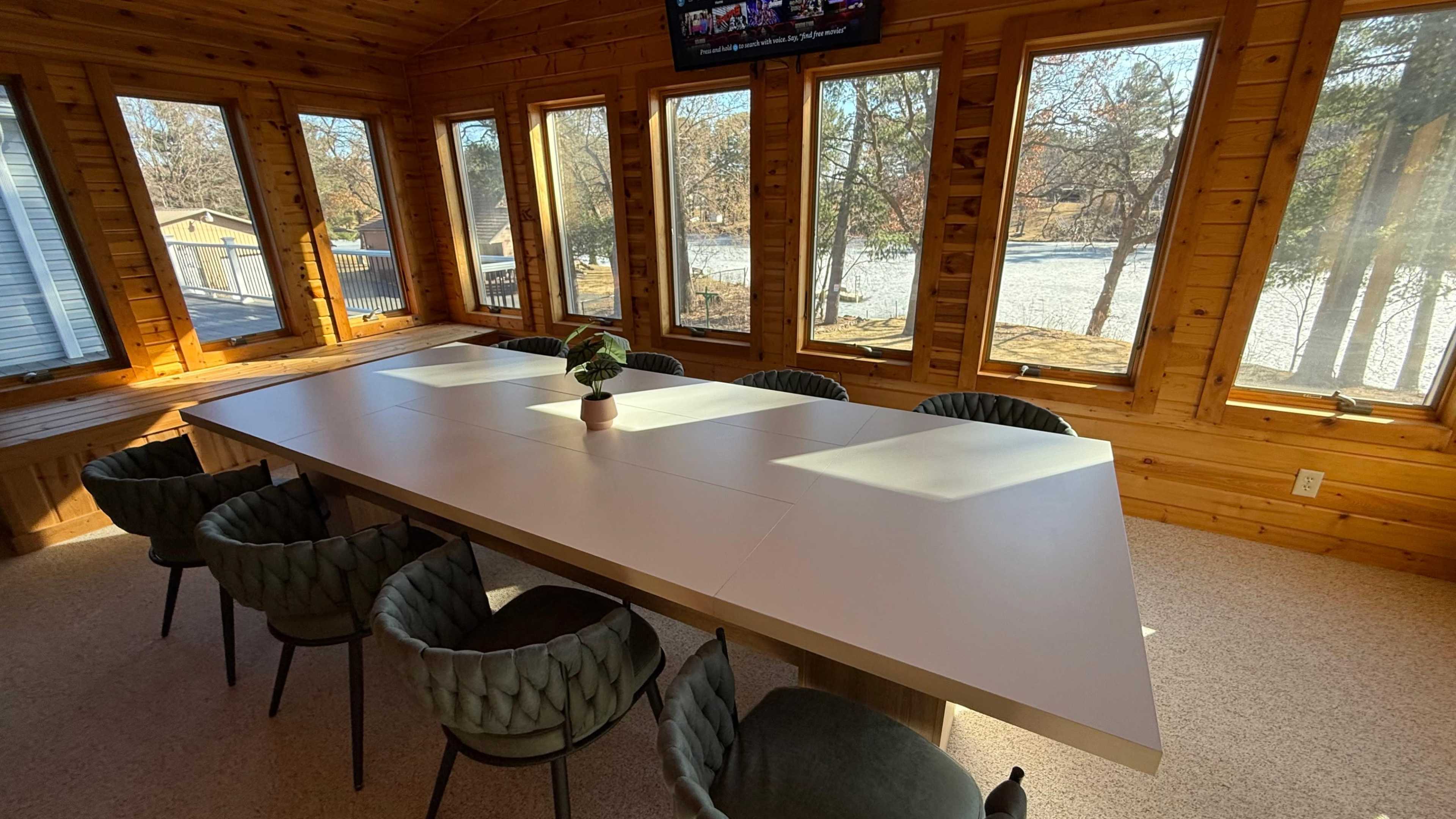 The image shows a spacious conference room with a long white table surrounded by eight chairs and large windows providing a view of an outdoor landscape.