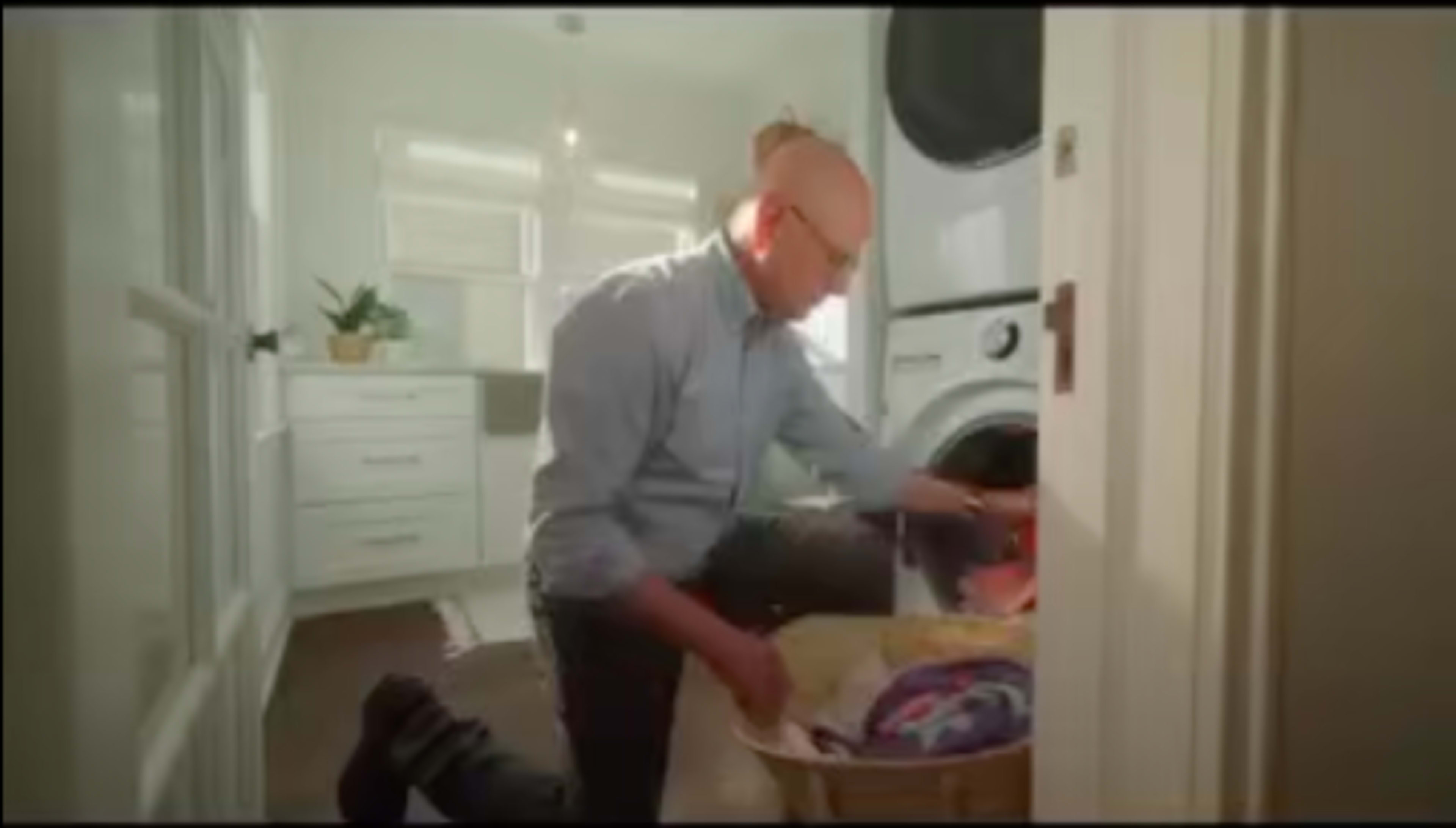 A man in a collared shirt is kneeling next to a laundry basket while removing clothes from a washing machine in a bright laundry room.