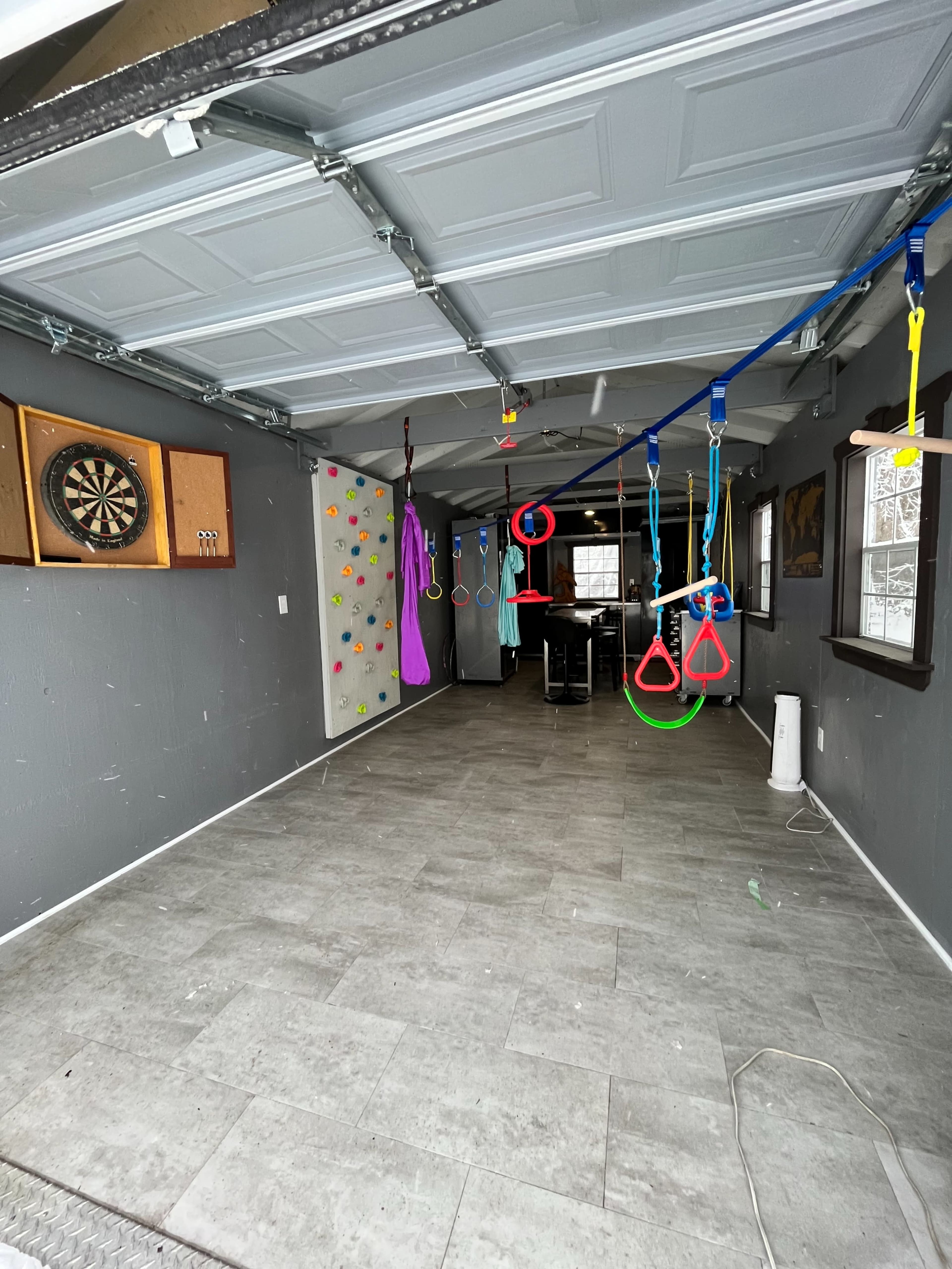 A garage space with a climbing wall, a dartboard mounted on the wall, and various gymnastic rings hanging from the ceiling.