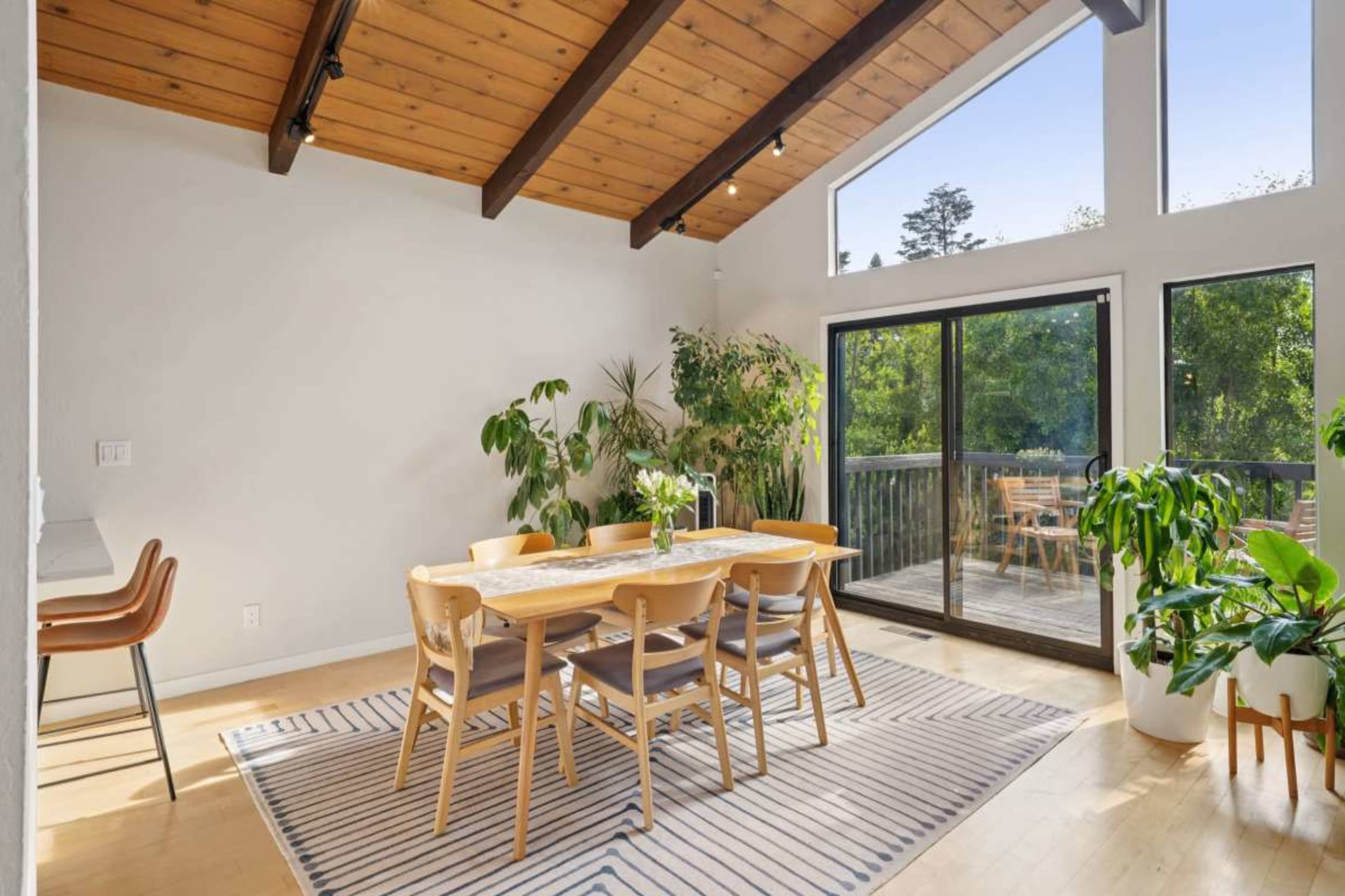 A spacious dining area features a wooden table surrounded by chairs, large windows, and potted plants, with a sliding glass door leading to an outdoor deck.