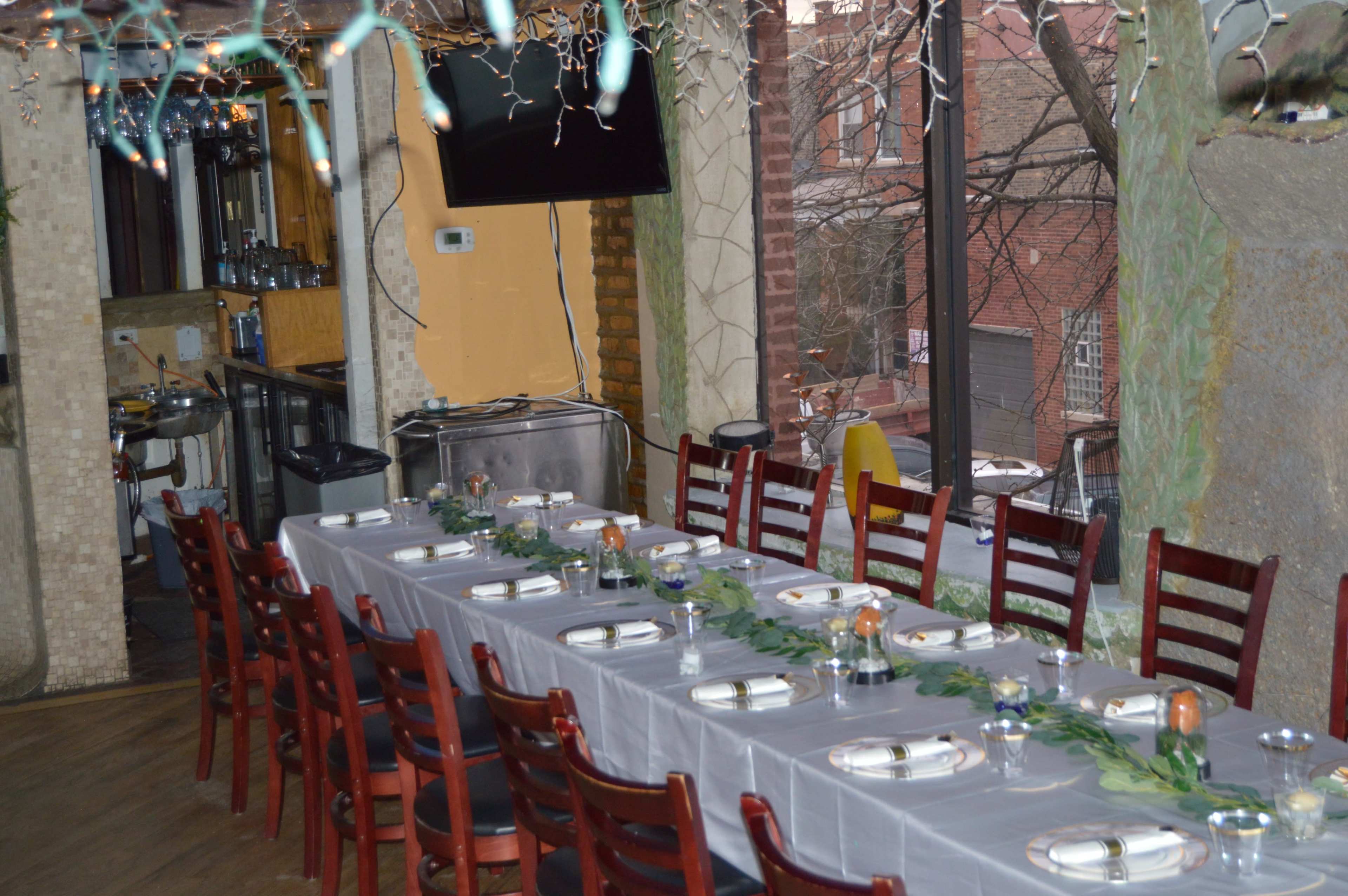 A long dining table is set for a meal, with plates, glasses, and decorative elements, in a restaurant with exposed brick and large windows.