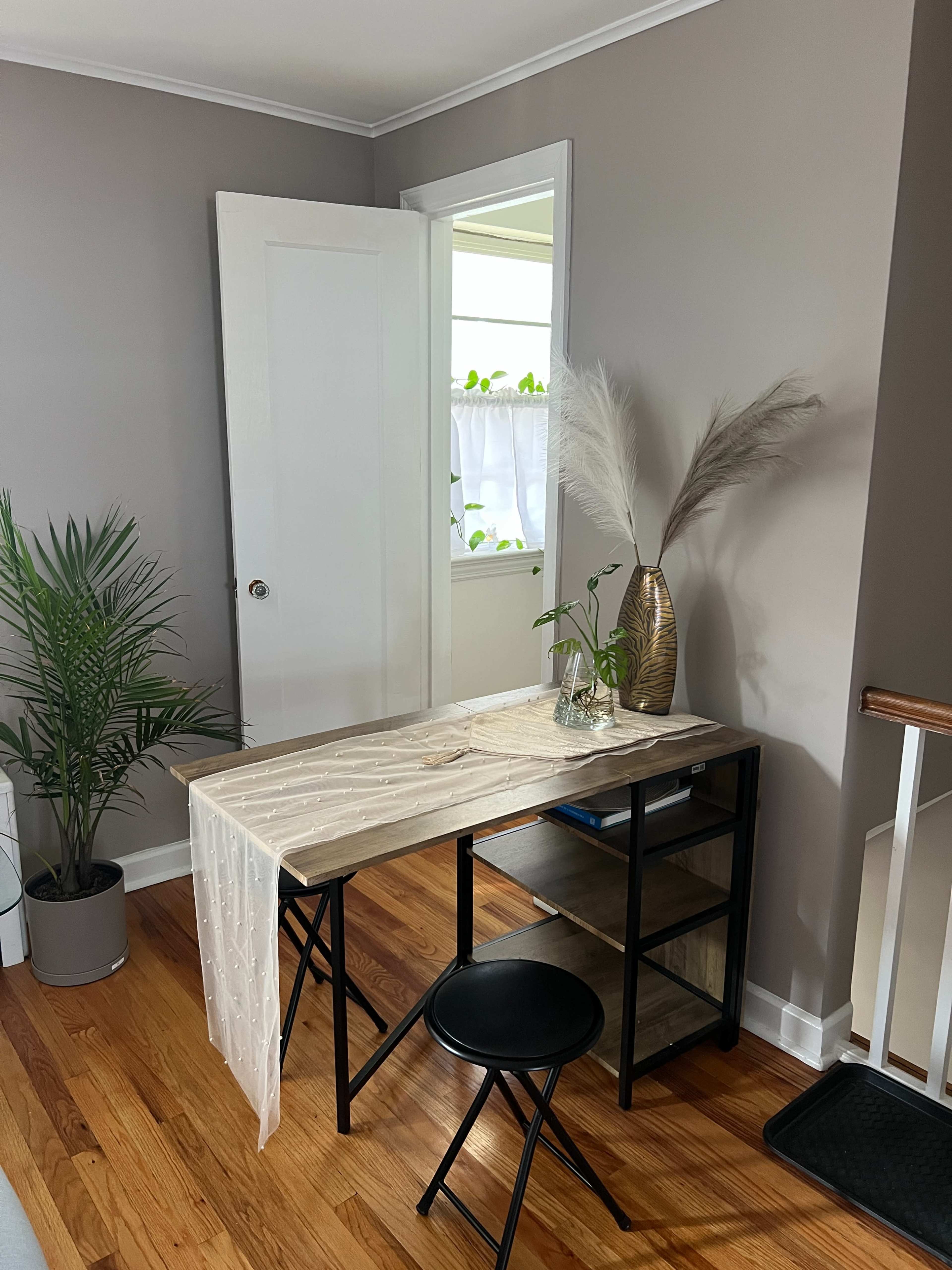 The image shows a small home office area featuring a wooden desk with a tablecloth, a black stool, a plant in a pot, and a decorative vase with pampas grass against a wall painted in a neutral color.