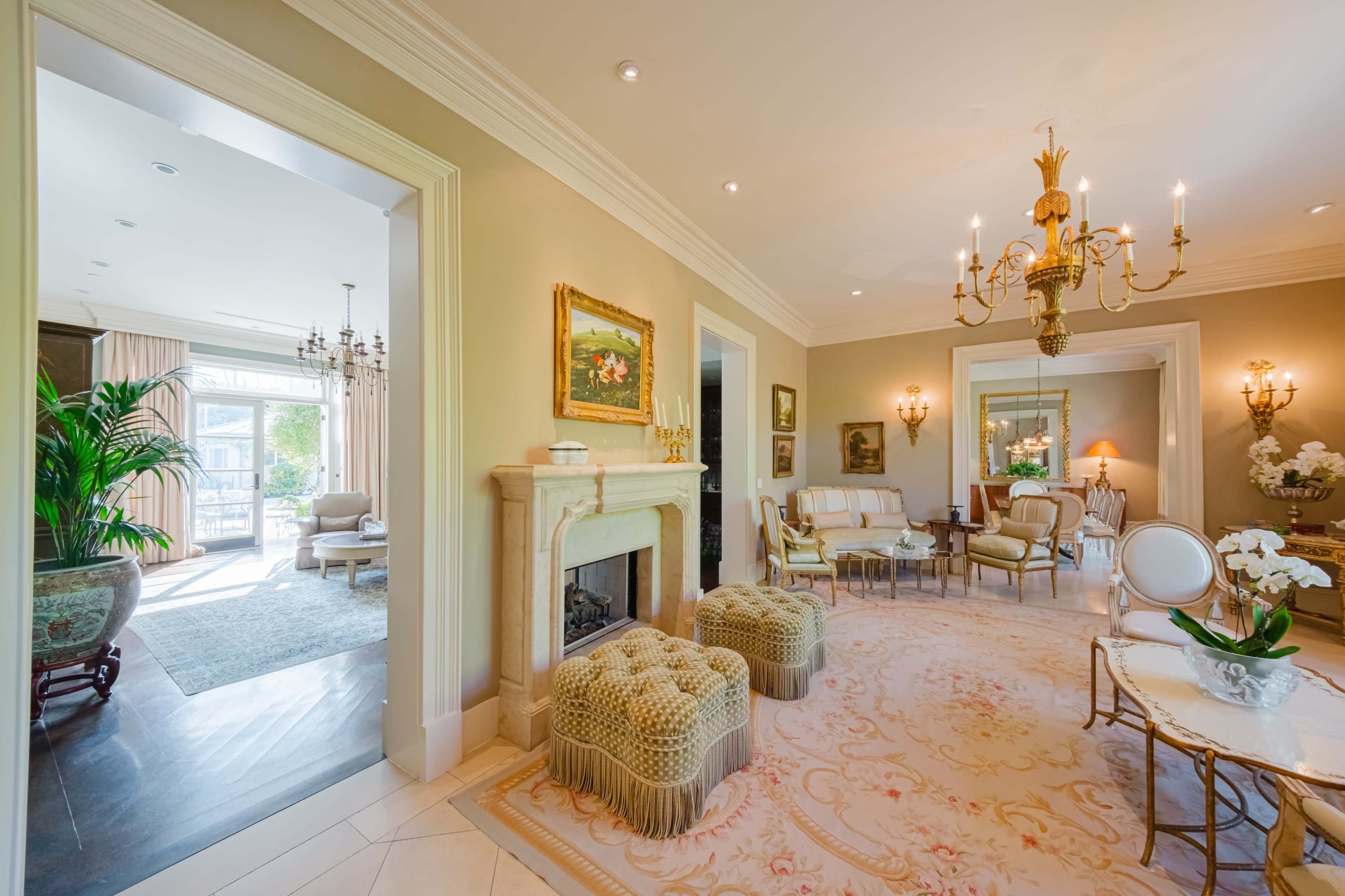 A bright and elegant living room features a chandelier, ornate furniture, and decorative rugs, with an adjoining space visible through an open doorway.