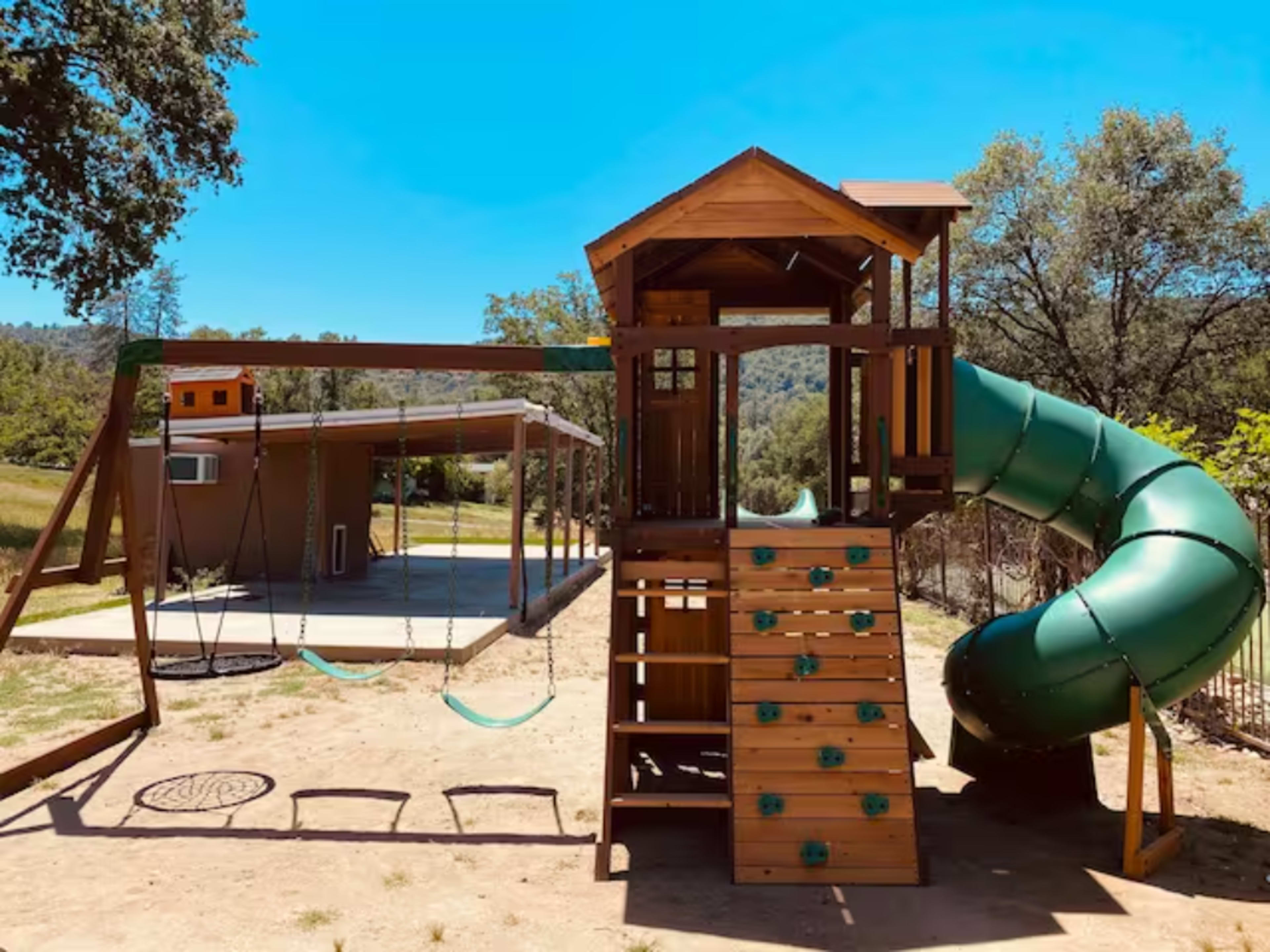 A wooden playground structure with a slide, climbing wall, and swings, set against a clear blue sky and green trees in the background.