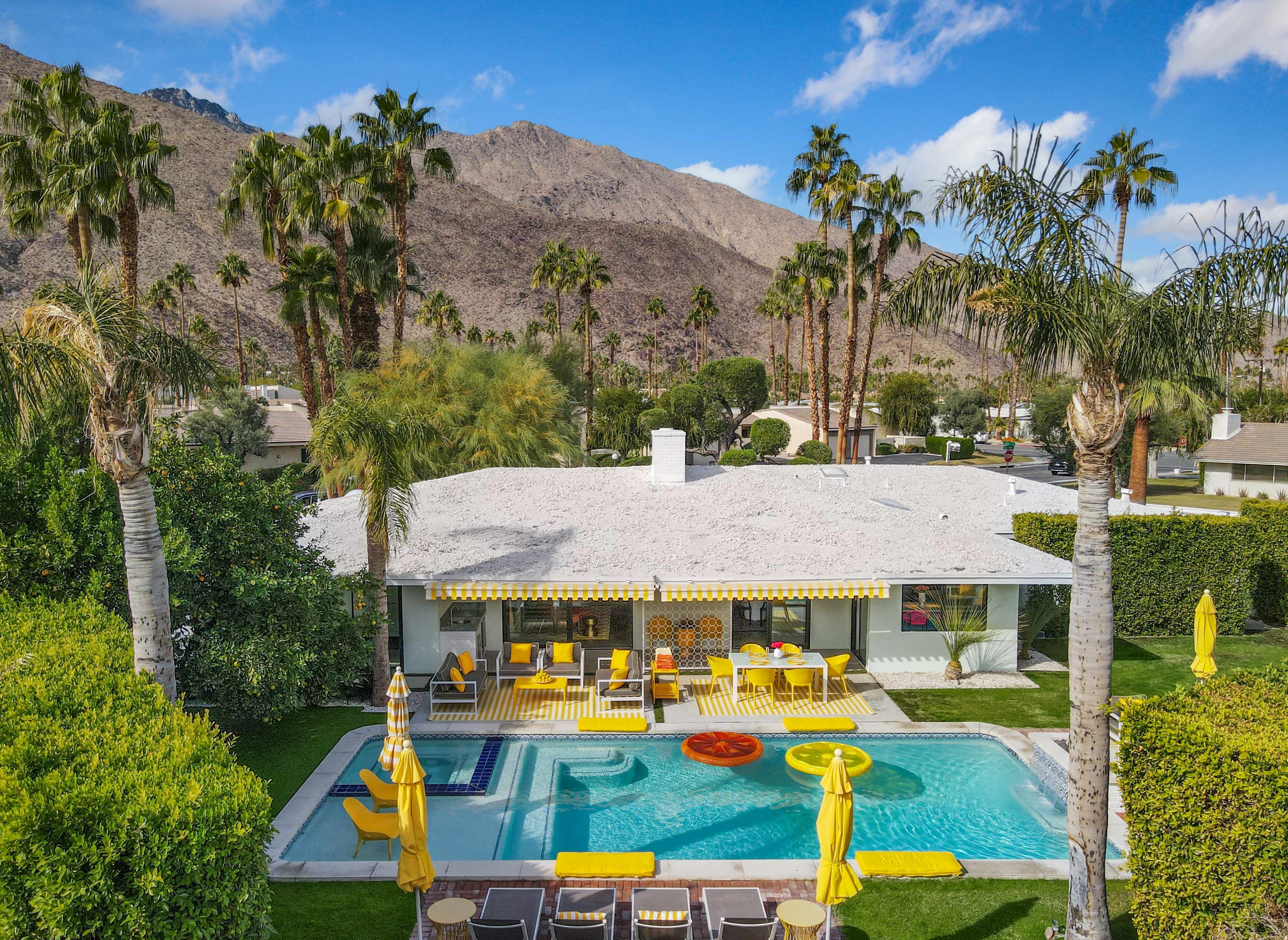 A modern home with a yellow-themed outdoor area surrounds a swimming pool, set against a backdrop of mountains and palm trees.