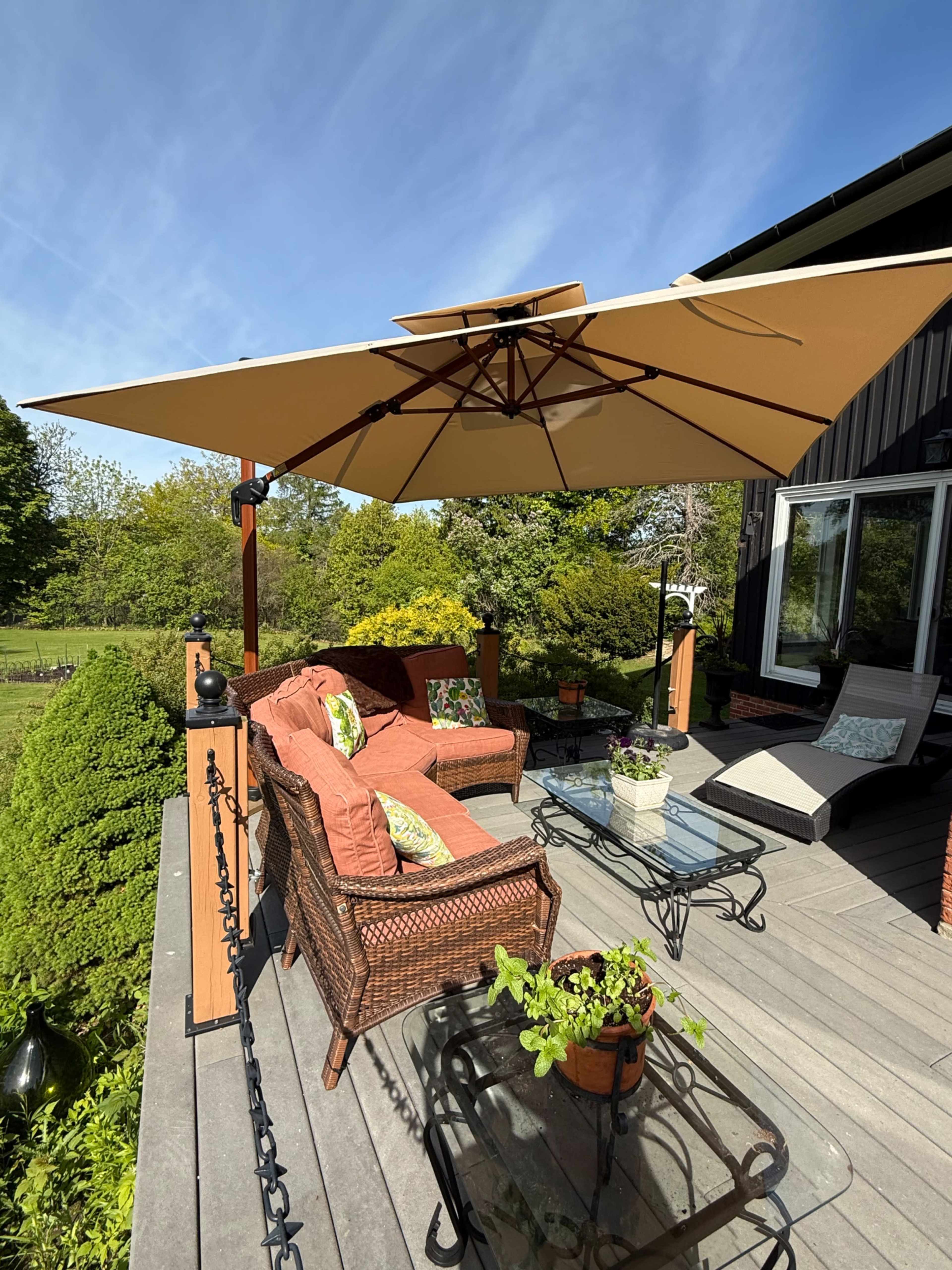 A patio features a set of wicker furniture, including a sofa and chairs, under a large umbrella, surrounded by greenery.