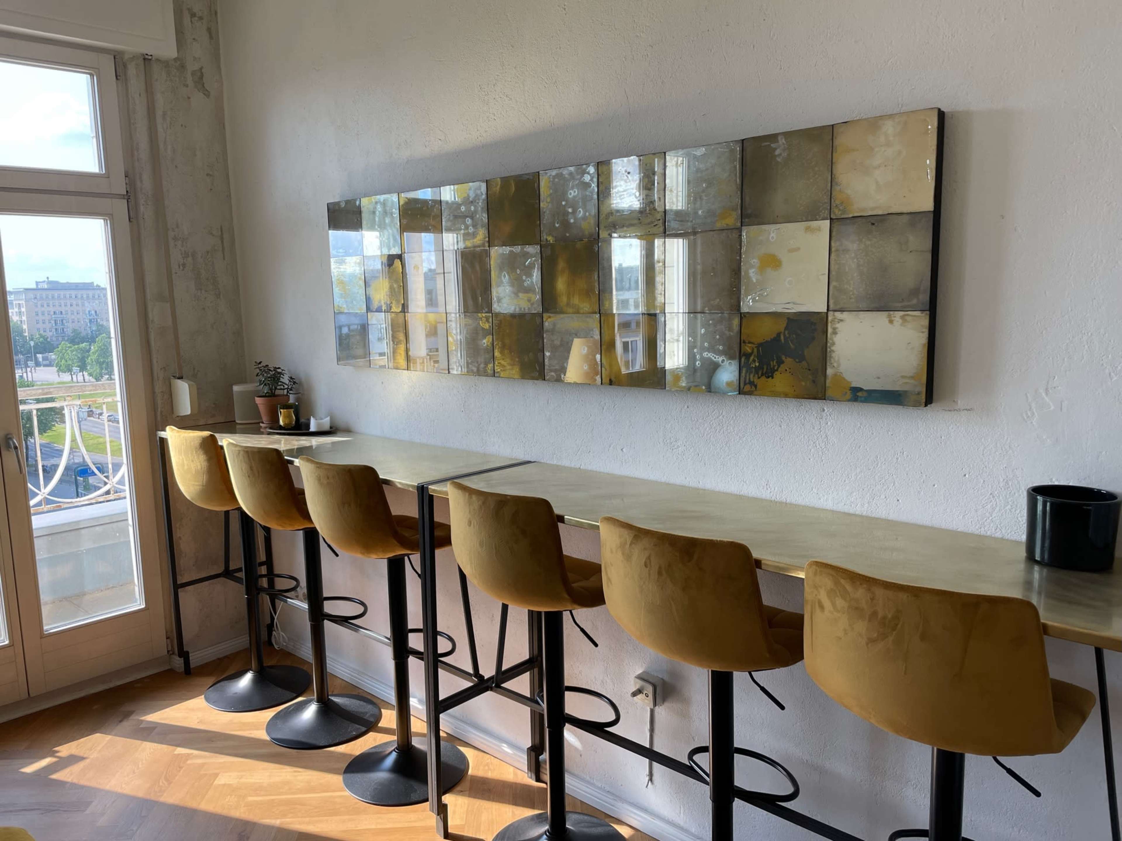A modern cafe interior with a row of yellow upholstered bar stools facing a long, slender table beneath a decorative wall mirror composed of square panels.