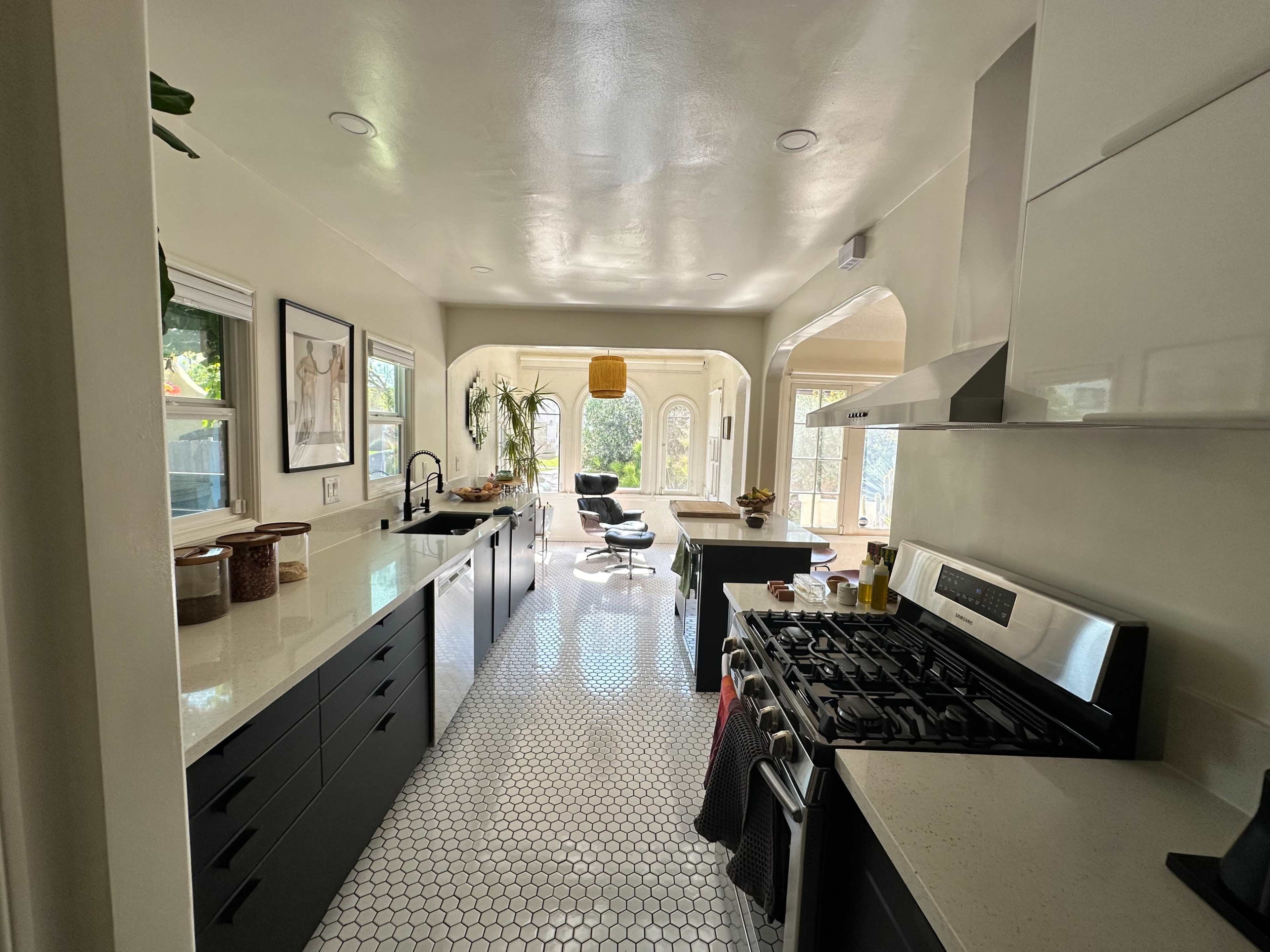 A modern kitchen features sleek black cabinetry, a stainless steel stove, and a hexagonal tile floor, leading to a brightly lit dining area with large windows.