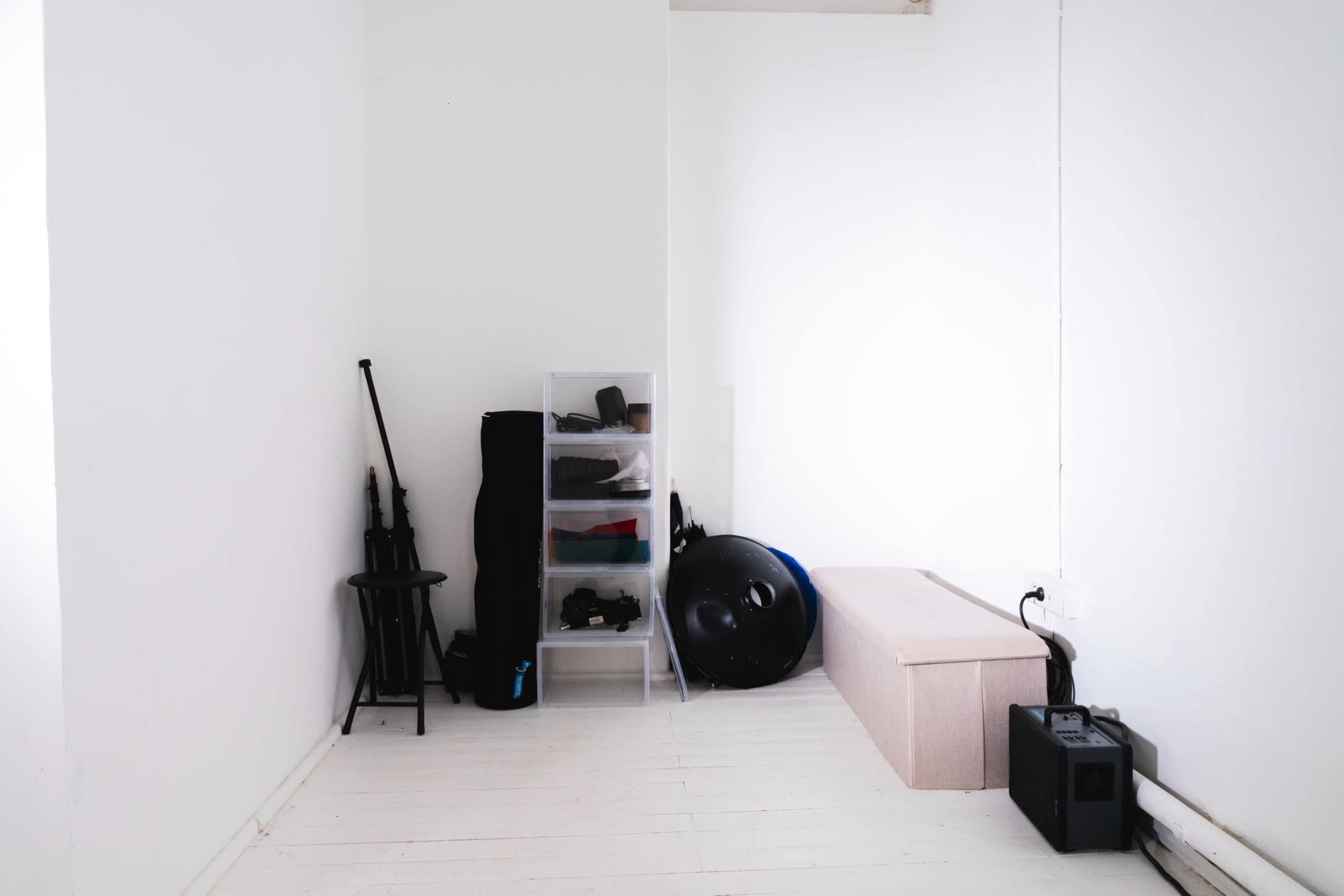 The image shows a minimalistic room with white walls, featuring a storage unit with various items, a black stool, a large exercise ball, and a low bench.