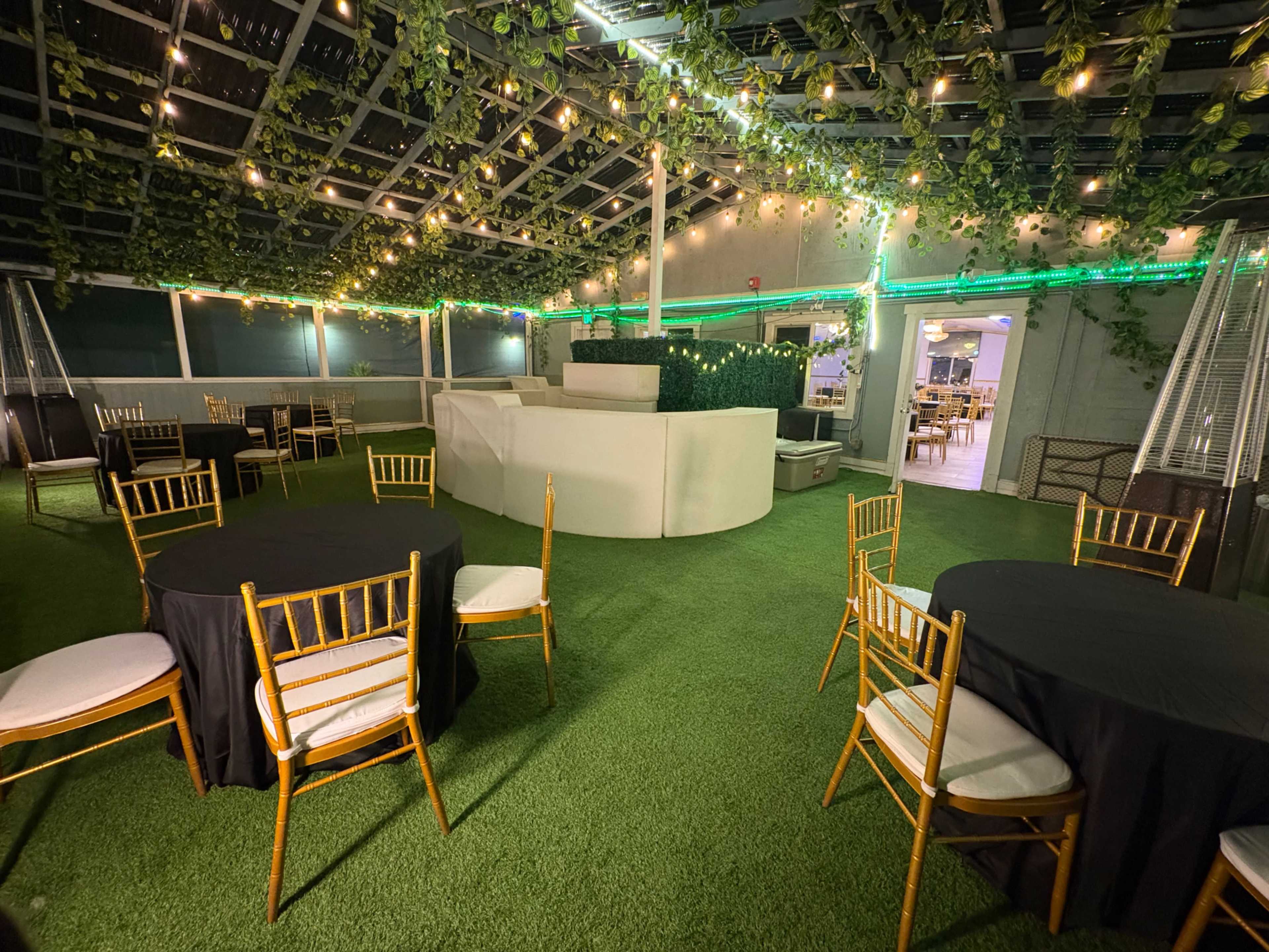 The image shows a rooftop venue with several round tables covered in black and white tablecloths, surrounded by greenery and string lights.