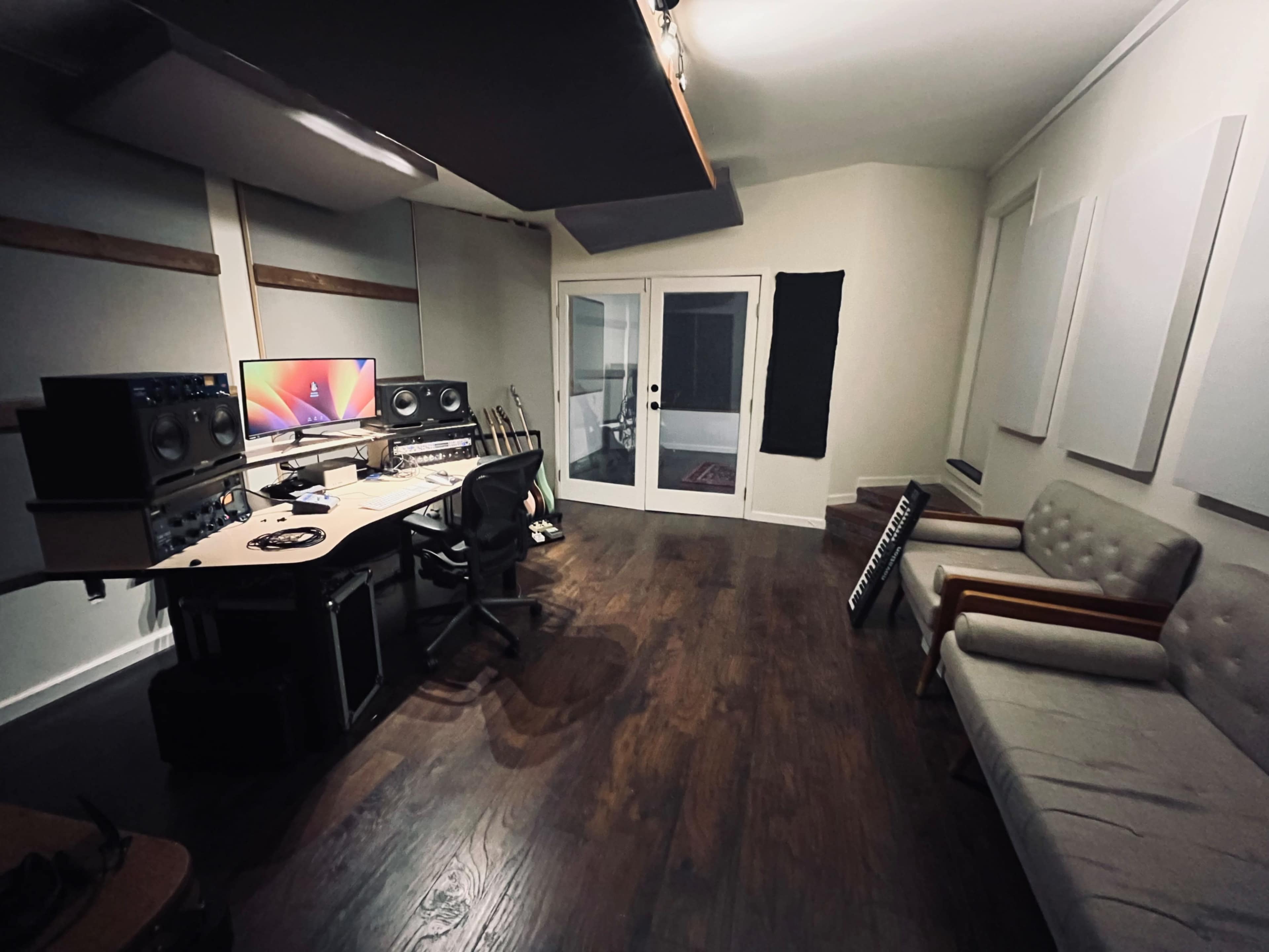 A music studio featuring a mixing desk with speakers and a computer setup, a sofa, and a glass door leading to an outdoor area.