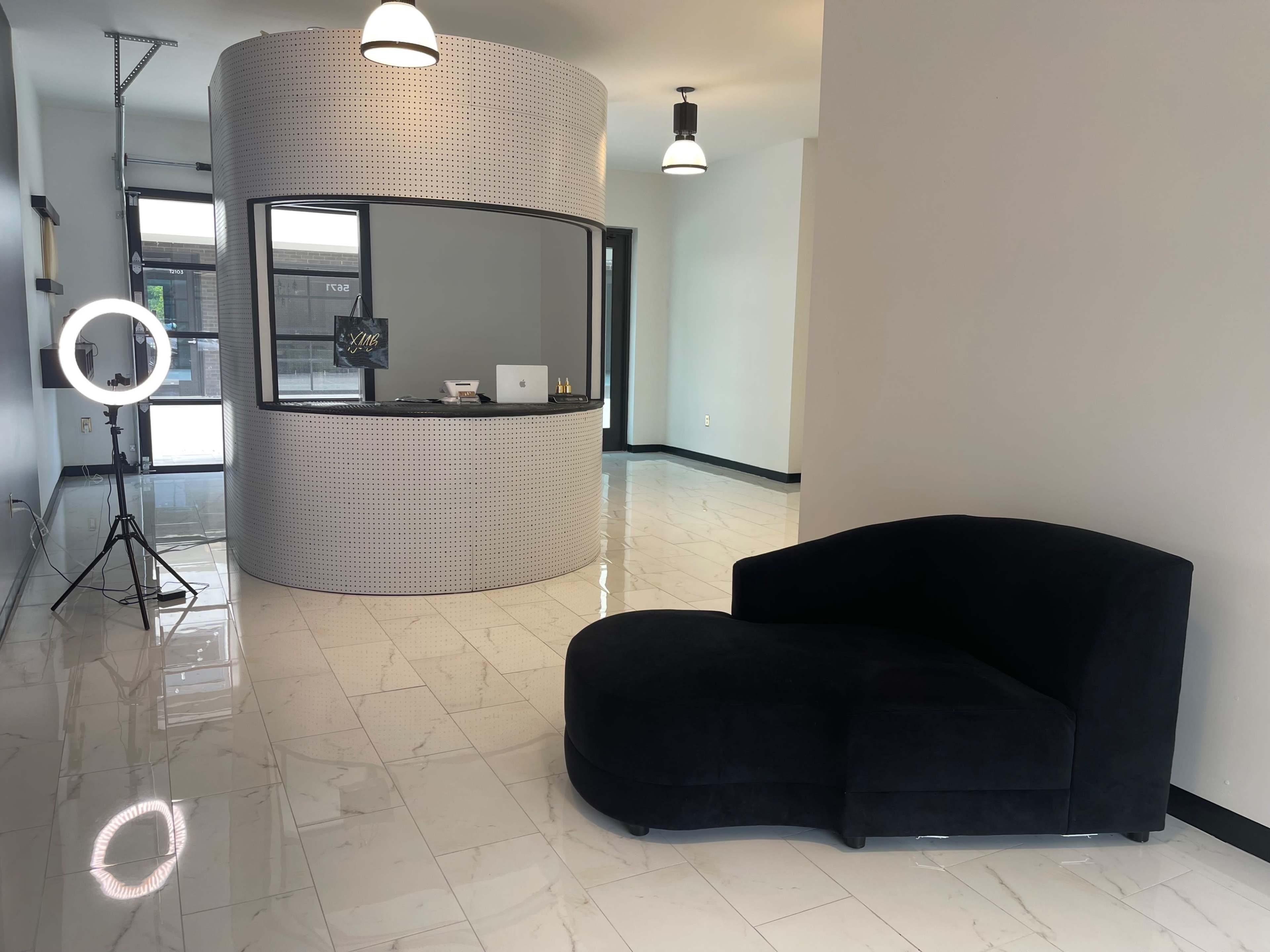 The image shows a modern interior space featuring a curved reception desk, a black chaise lounge, and a ring light setup on a polished tiled floor.