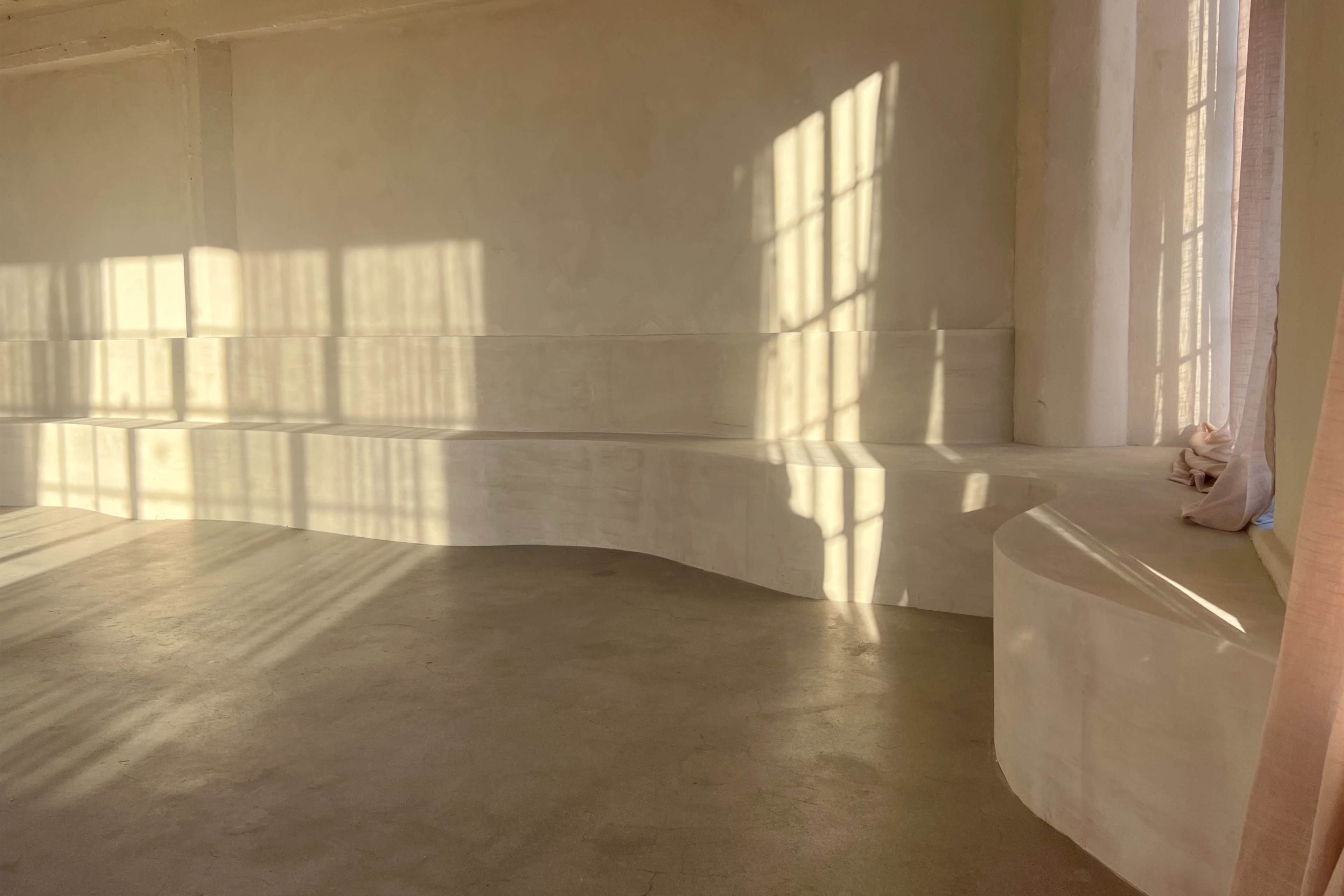 A spacious, minimalistic room with curved white seating and sunlight casting shadows through large windows.