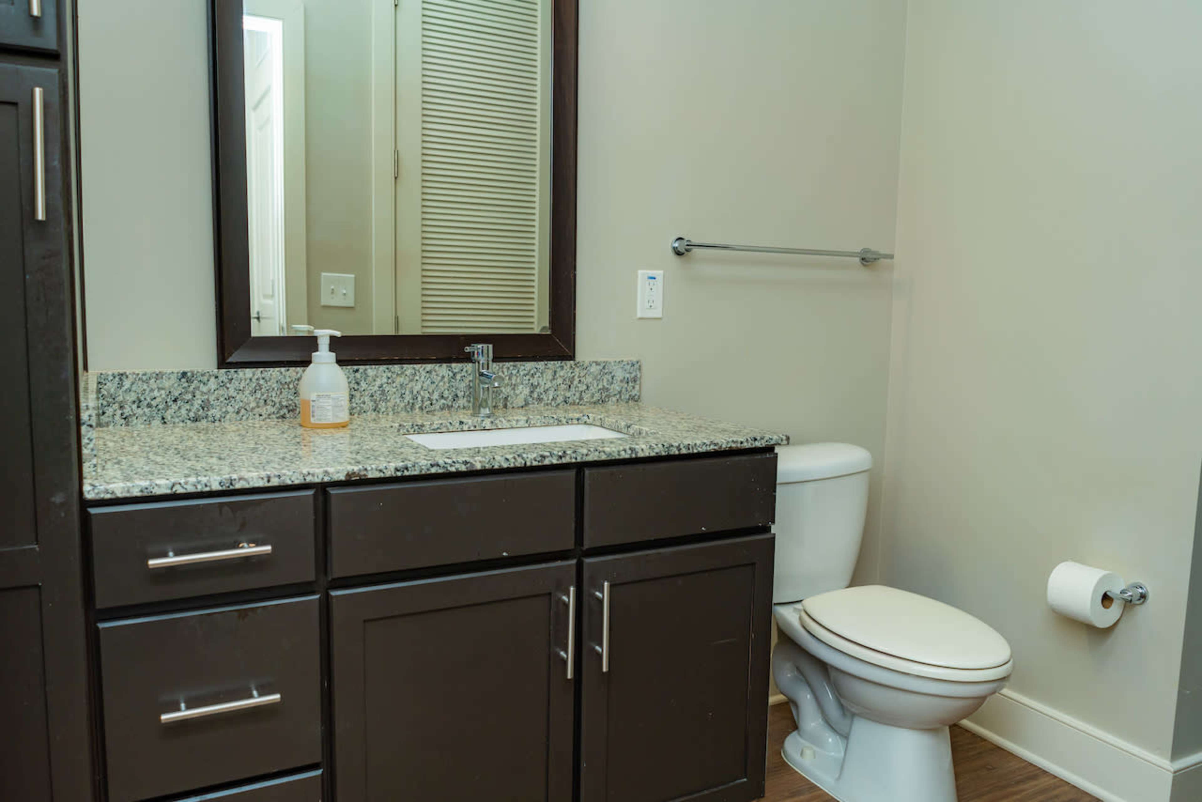 A bathroom with a dark cabinetry, a granite countertop, a mirror, a toilet, and a towel bar.