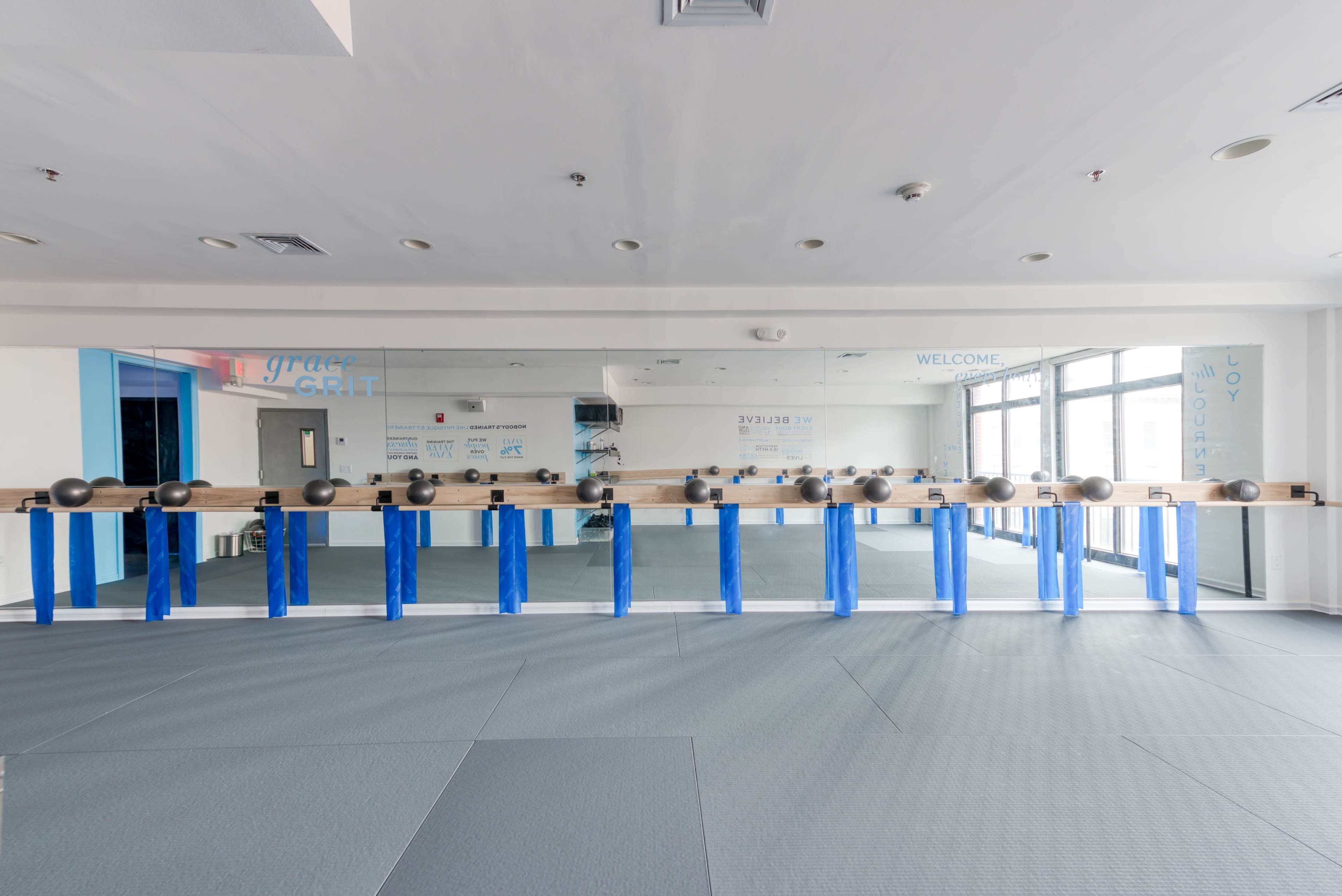 The image shows a spacious, bright exercise studio with a mirrored wall and a row of weights on a wooden bar, situated above gray floor mats.