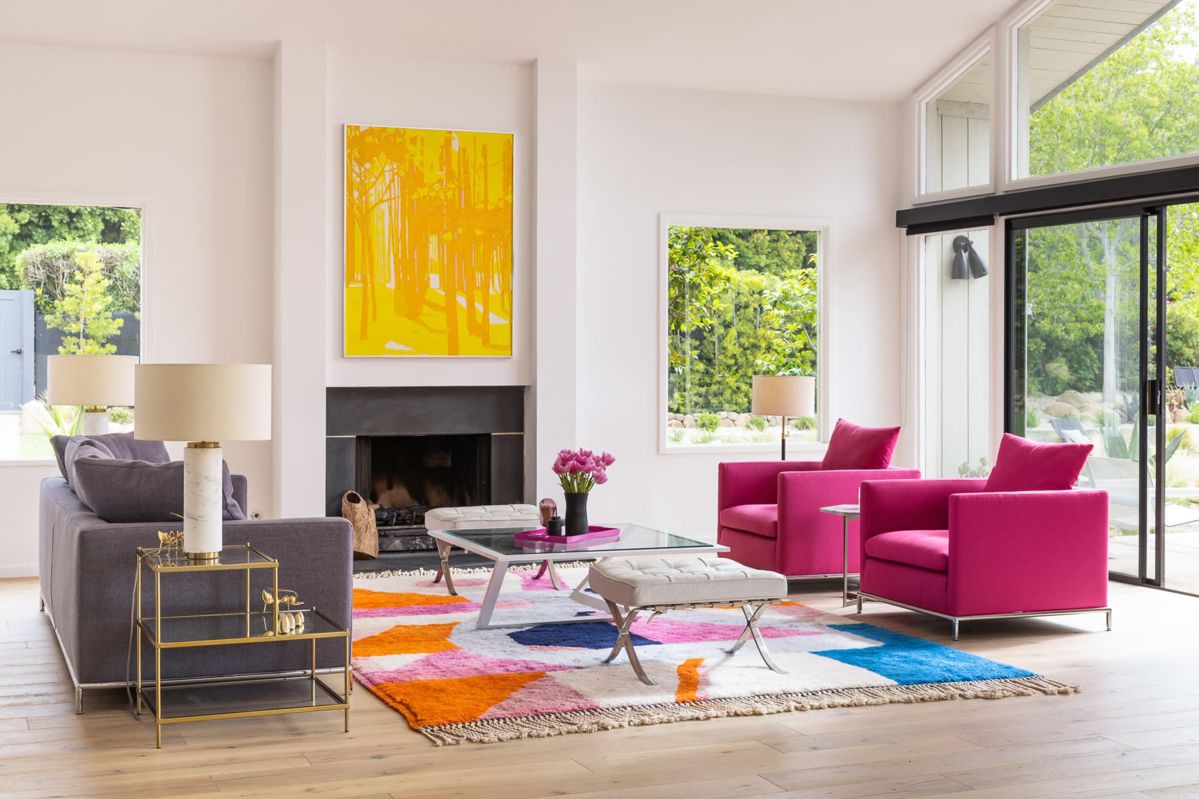 The living room features modern furniture, including a gray sofa and bright pink armchairs, arranged around a glass coffee table with a colorful area rug and a large yellow artwork on the wall.
