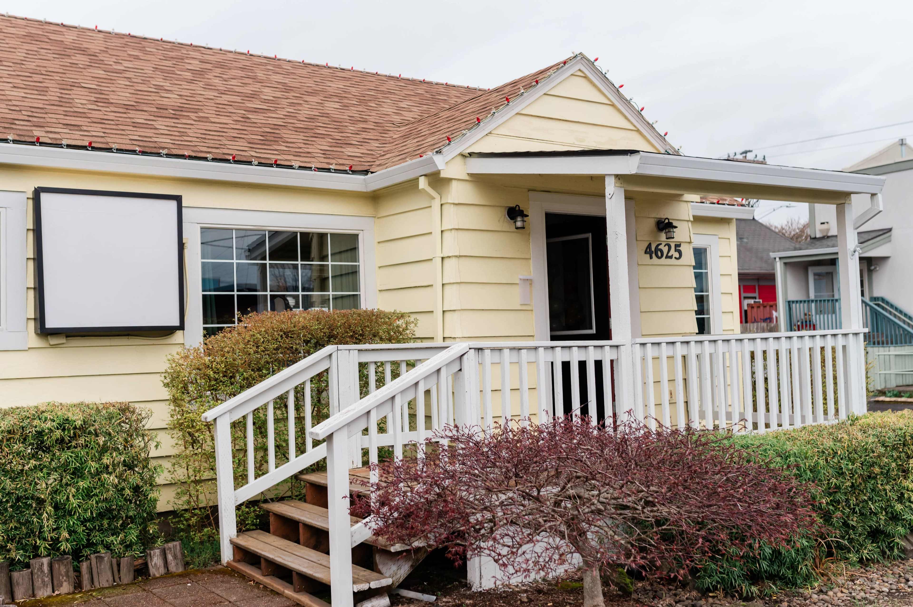 The image shows a single-story yellow house with a front porch, a sloped roof, and neatly trimmed landscaping.