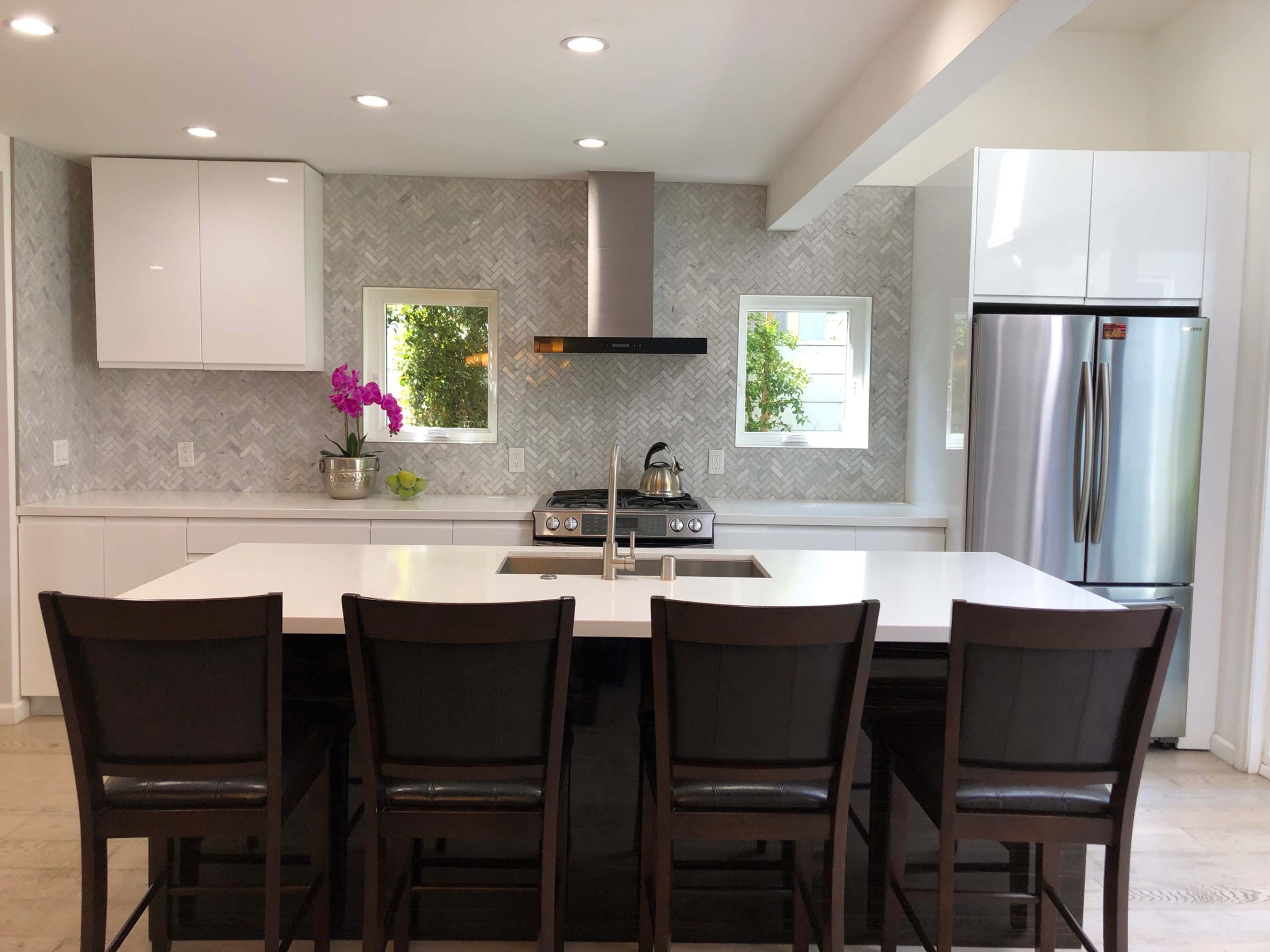 A modern kitchen features a large island with four dark wooden bar stools, stainless steel appliances, and a light-colored backsplash with a herringbone pattern.