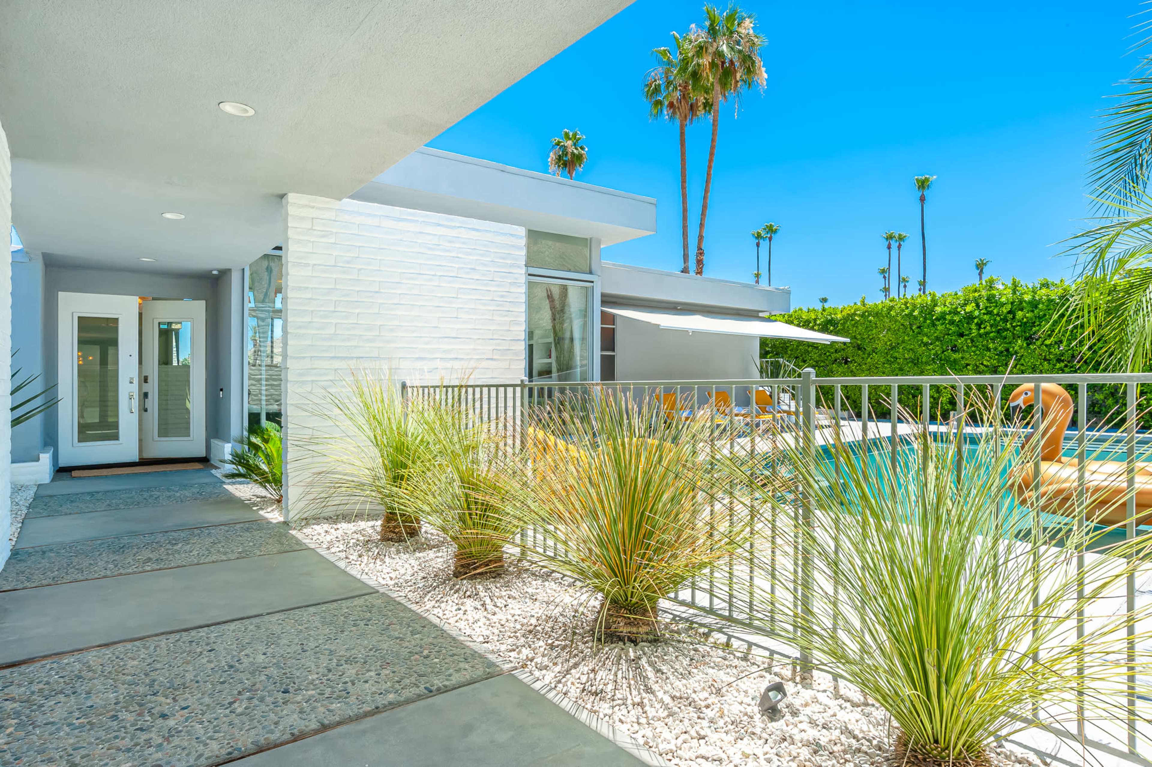A modern residential exterior featuring a pathway lined with gravel and plants, leading to a swimming pool surrounded by palm trees.