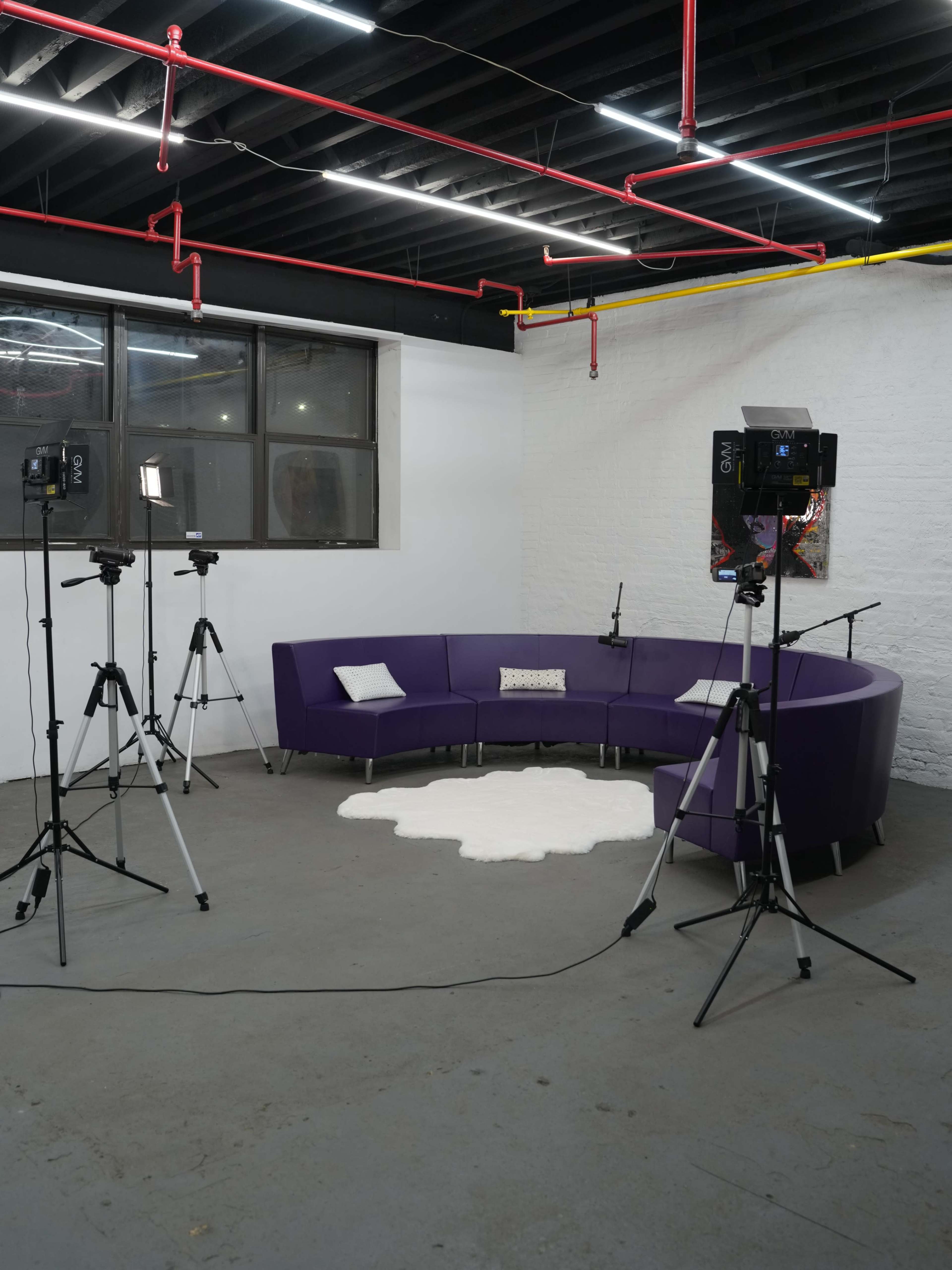 The image shows a minimalist studio with a purple circular couch, a white area rug, and three camera tripods positioned around the space.
