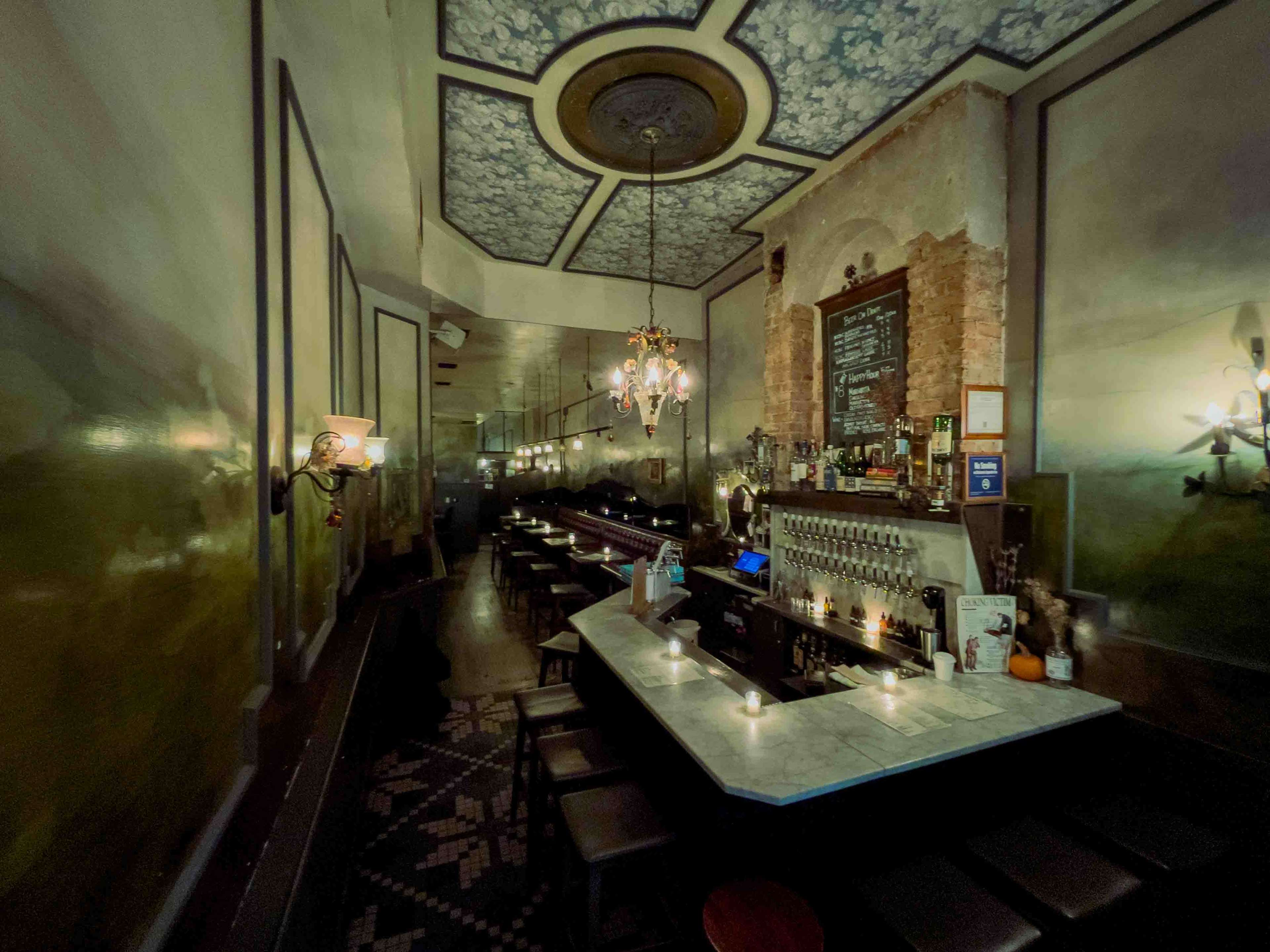 The image shows a dimly lit bar with a marble countertop, dark wooden seating, and vintage chandeliers, featuring murals on the walls and a counter displaying liquor bottles.
