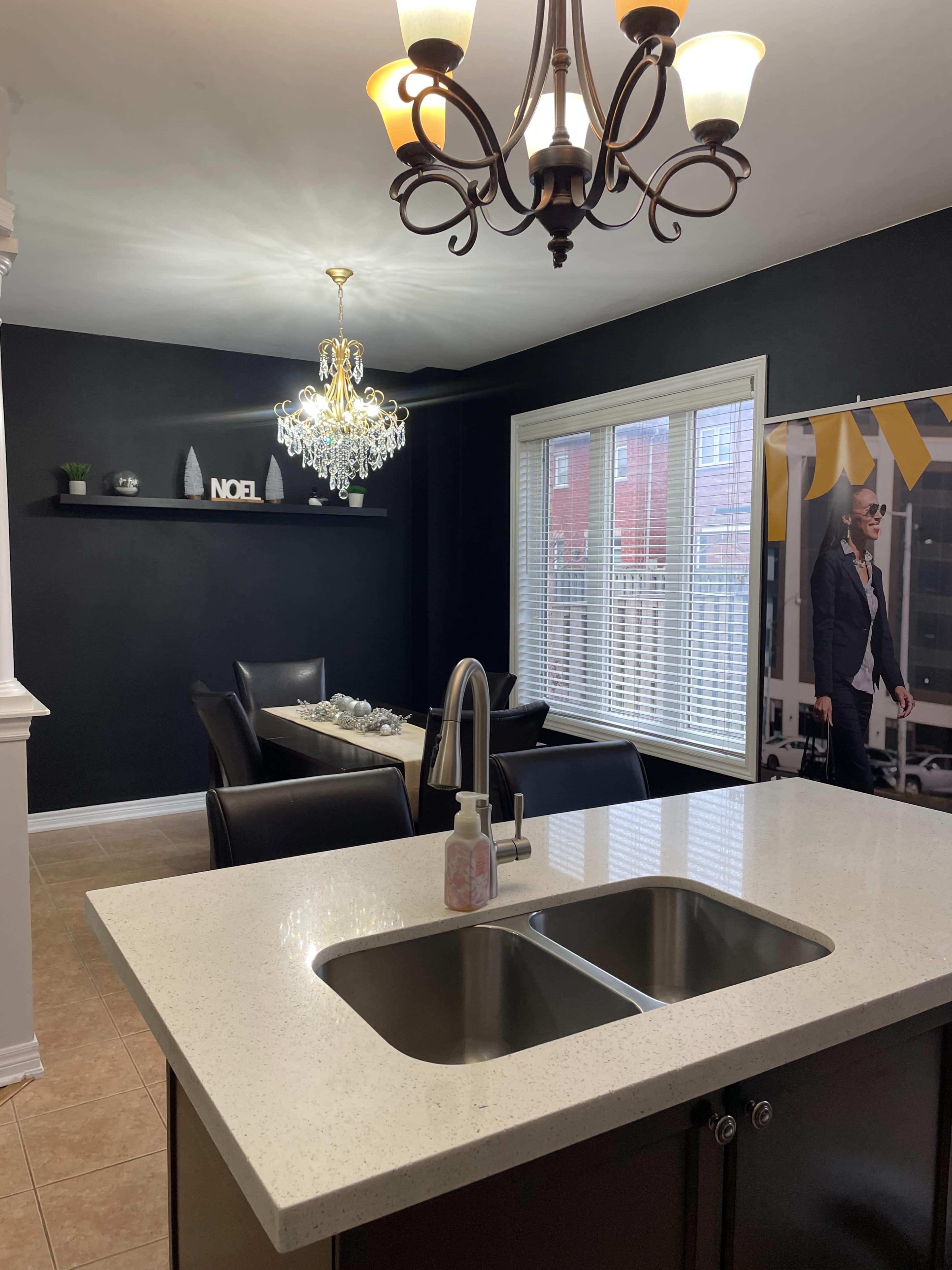 The image shows a modern kitchen and dining area featuring a double sink island, black walls, a chandelier, and a dining table with leather chairs.