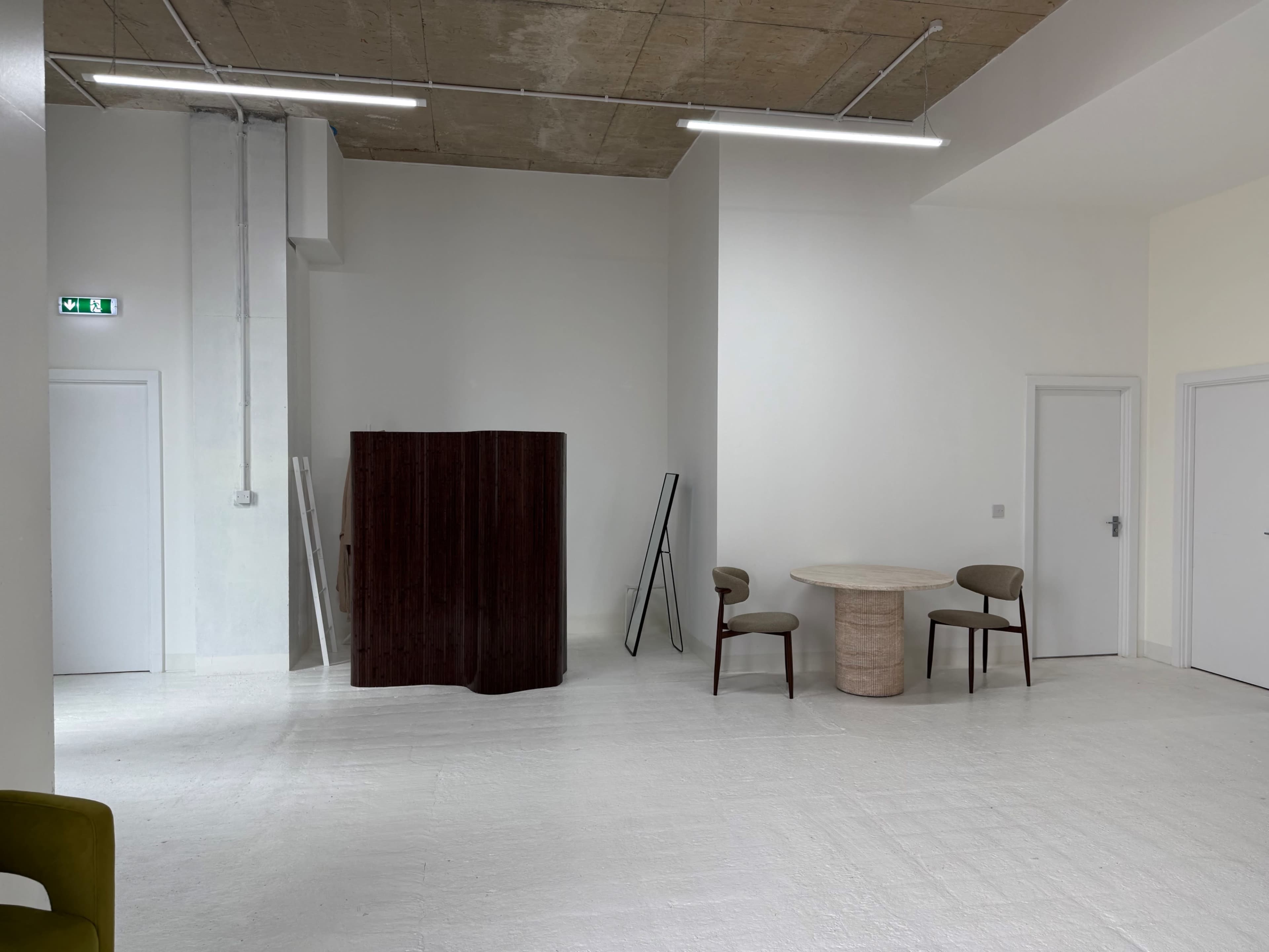 The image shows a minimalist interior space with a wooden screen, a round table, and two chairs, all set against white walls and a concrete ceiling.
