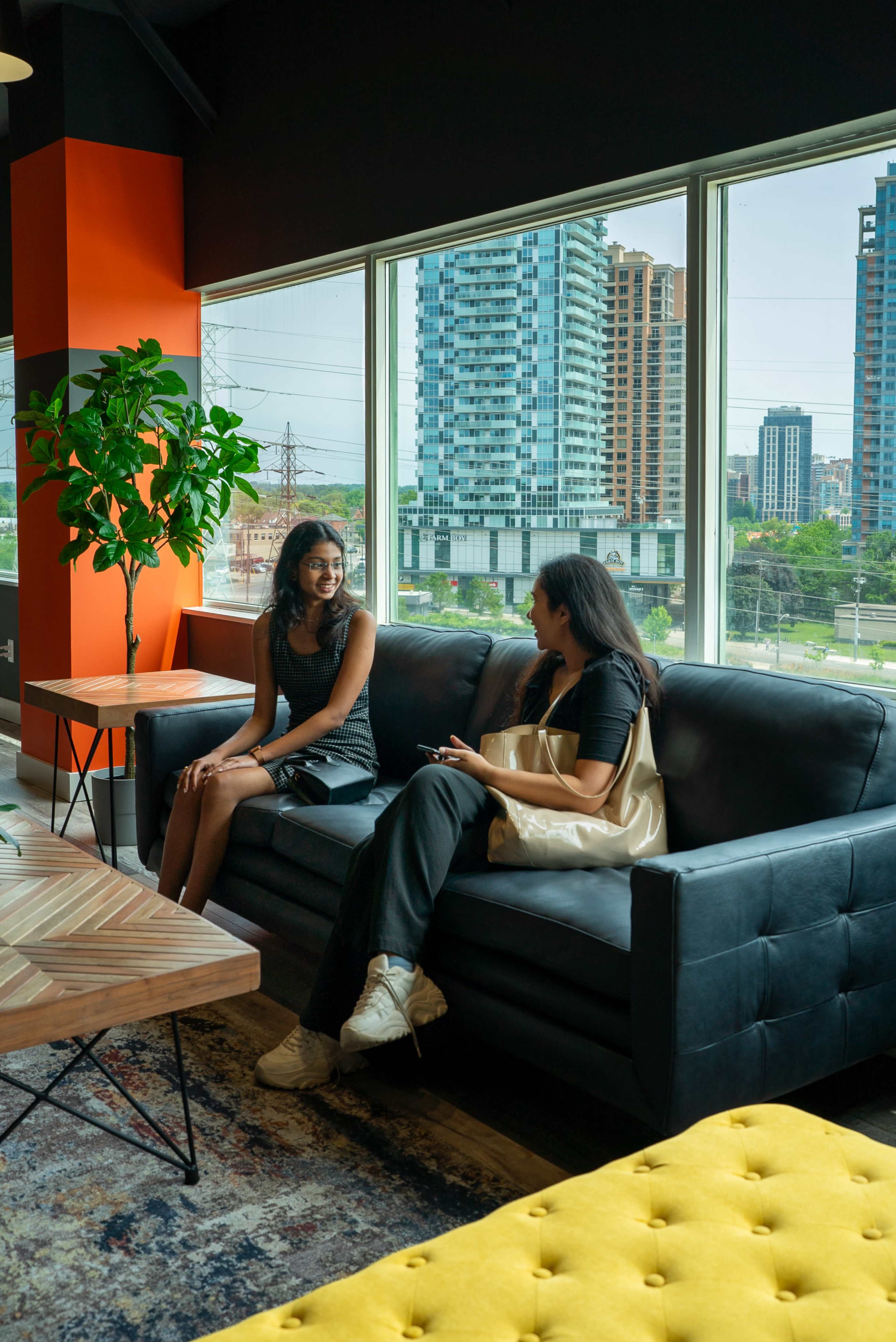 Two women are seated on a black sofa in a brightly lit room with large windows showcasing a view of urban buildings and greenery outside.