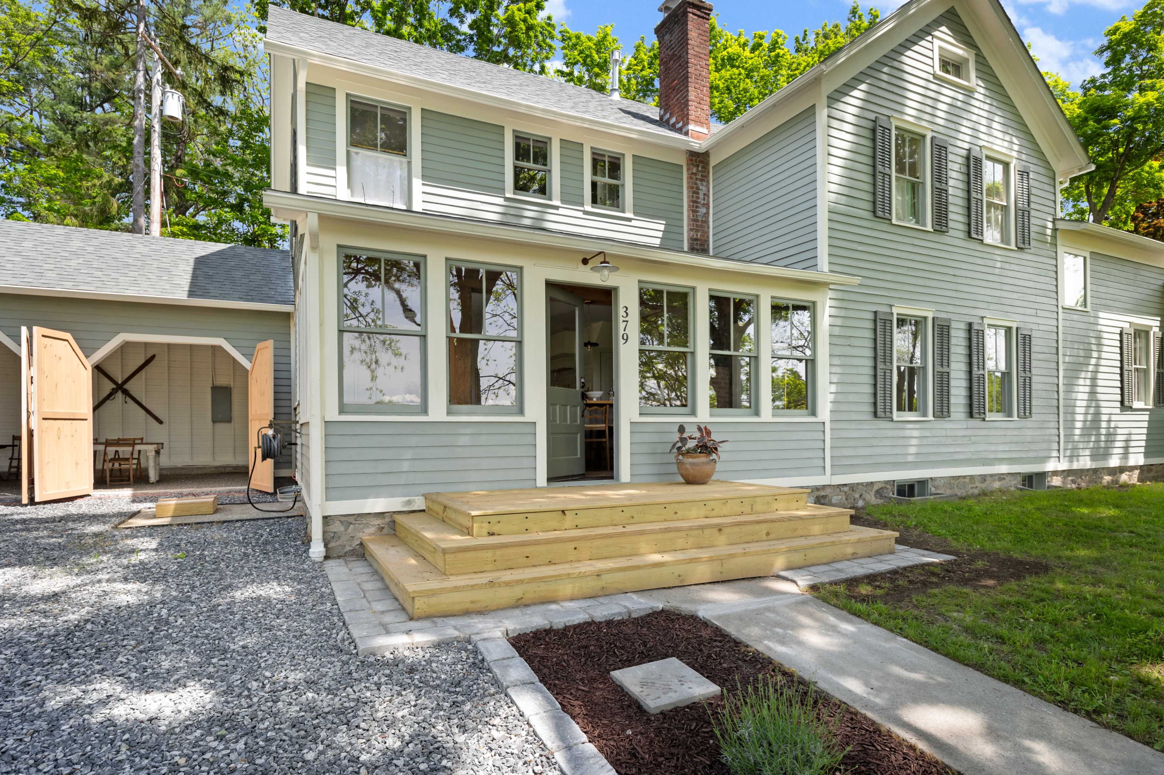 A light blue house with multiple windows, a wooden front porch, and a gravel pathway is set against a backdrop of trees.