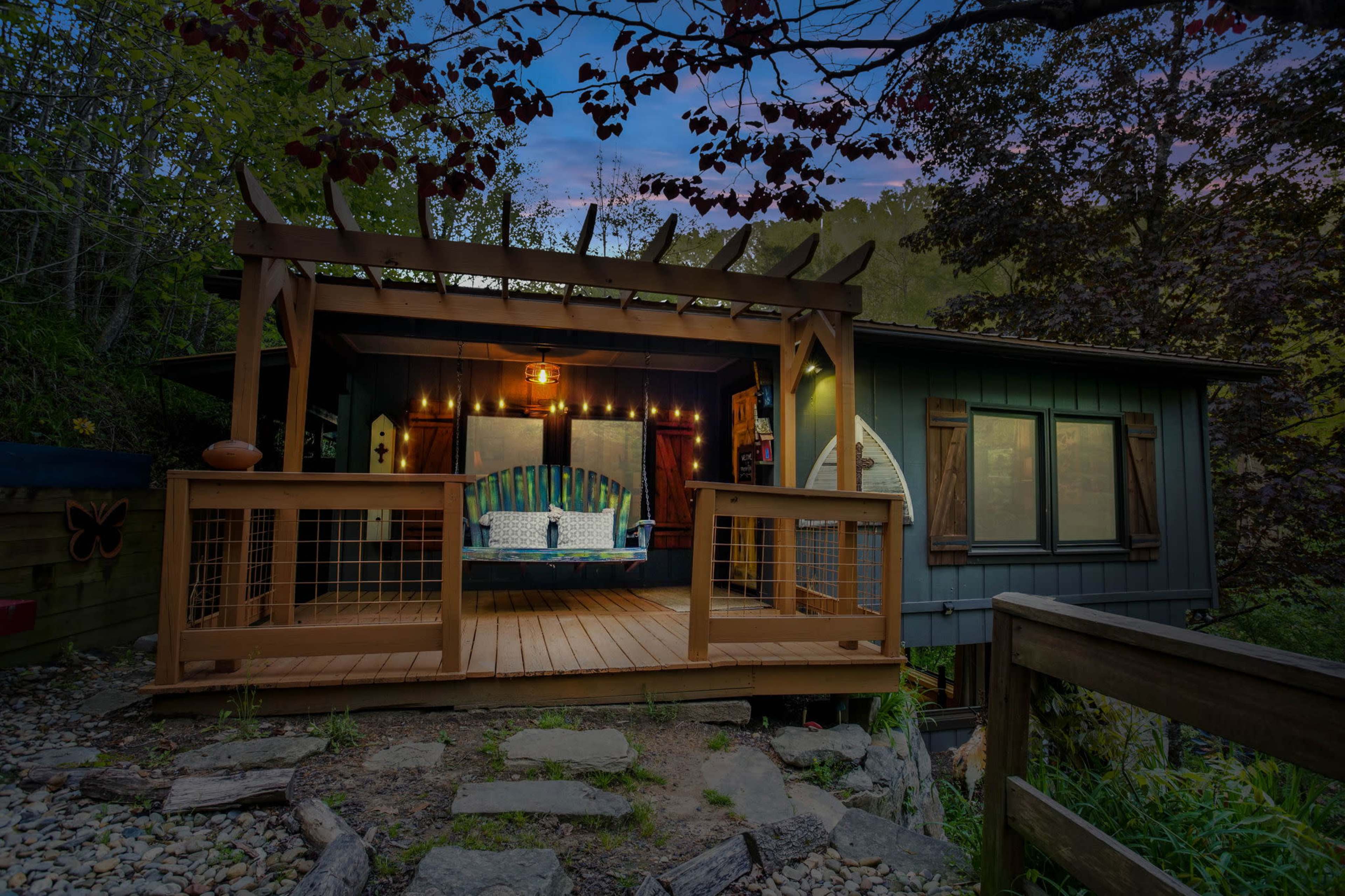 A wooden deck with a covered seating area is attached to a house surrounded by trees and rocks.