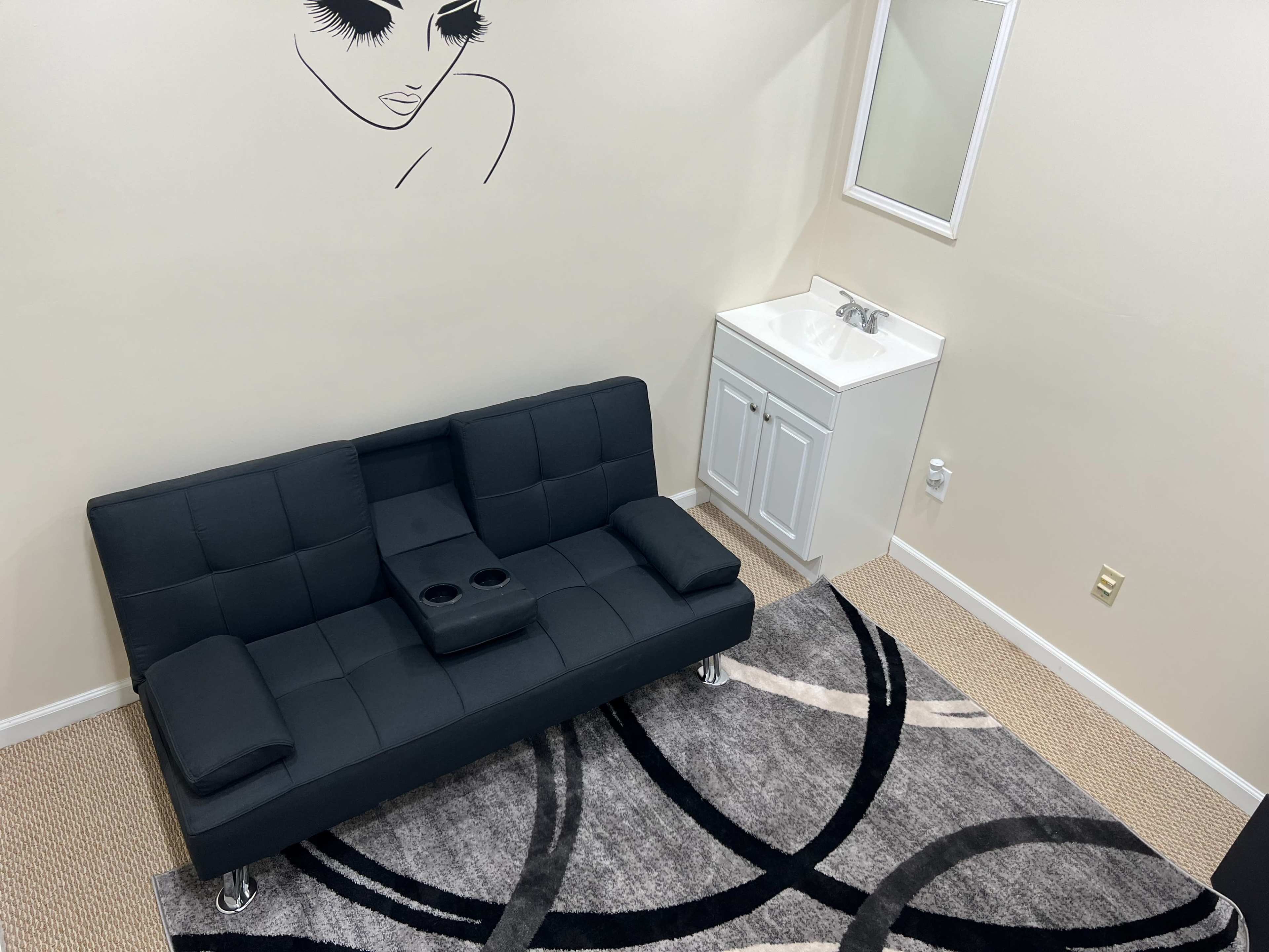 A black sofa with armrests sitting on a patterned rug in a room that has a sink and a wall decoration featuring an abstract face.