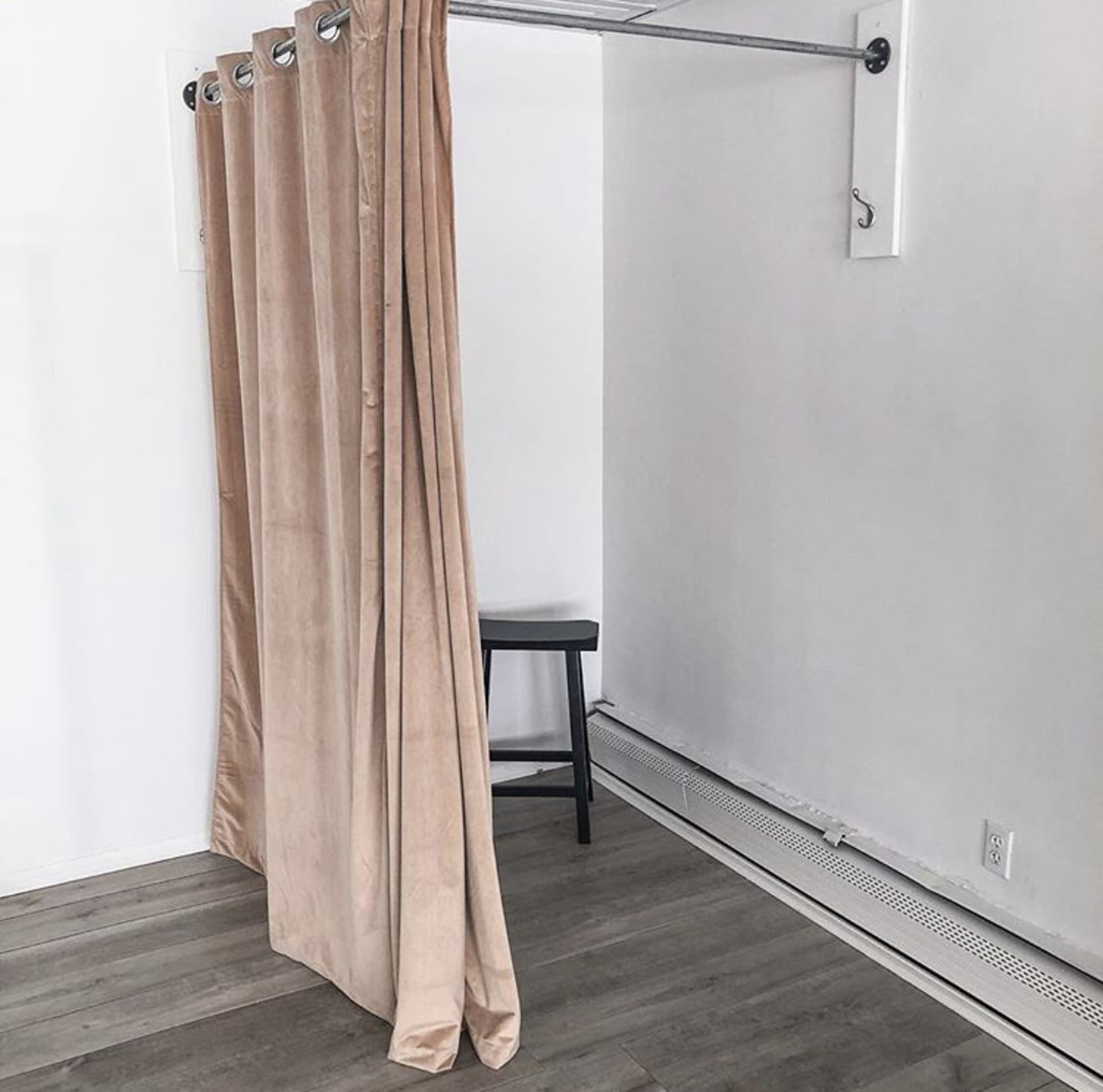 A set of beige curtains hangs from a rod in a room with wooden flooring and a stool nearby.
