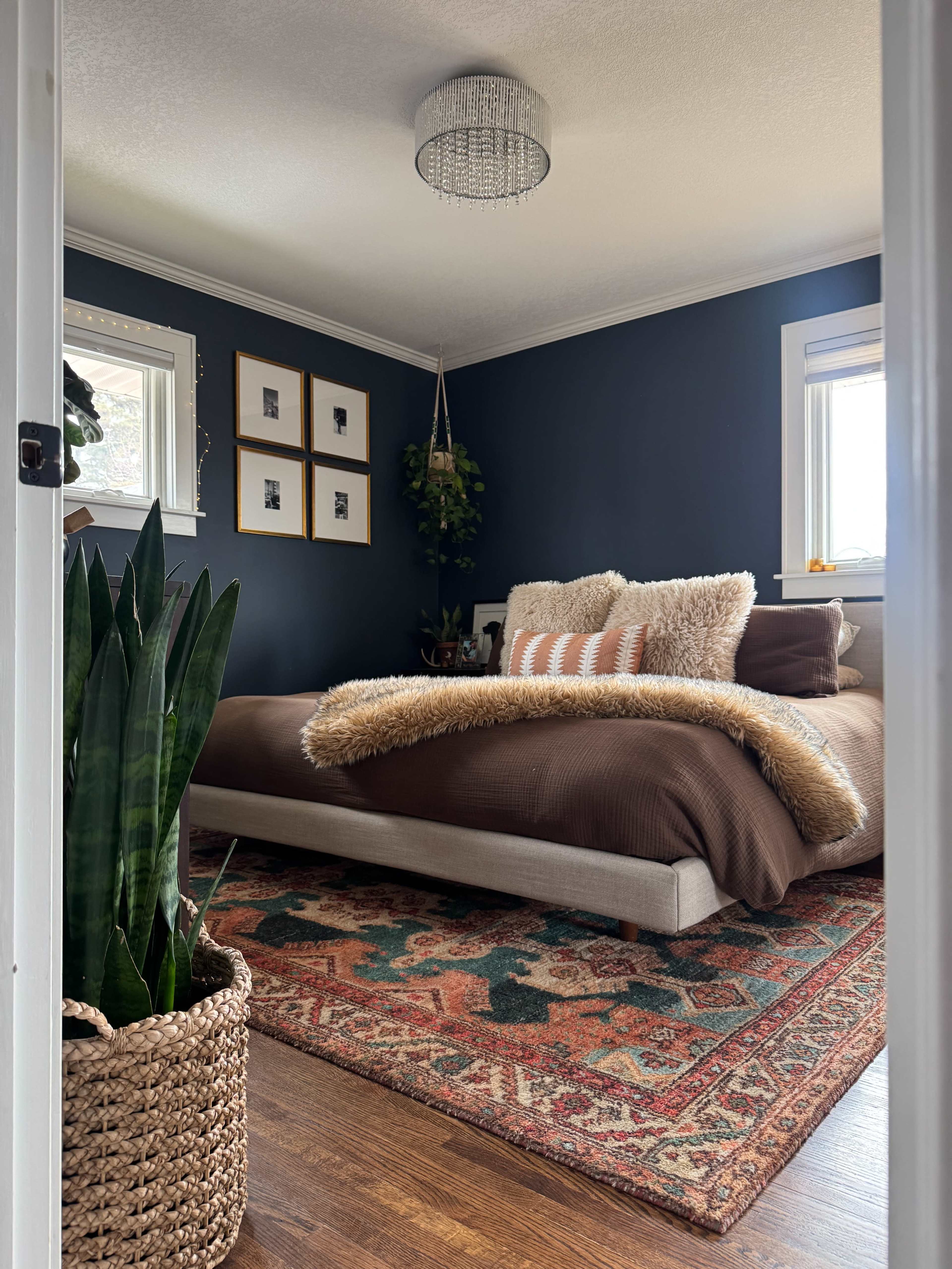 A cozy bedroom features a dark blue accent wall, a bed with a brown comforter, decorative pillows, a woven plant basket, and a vintage-style rug.